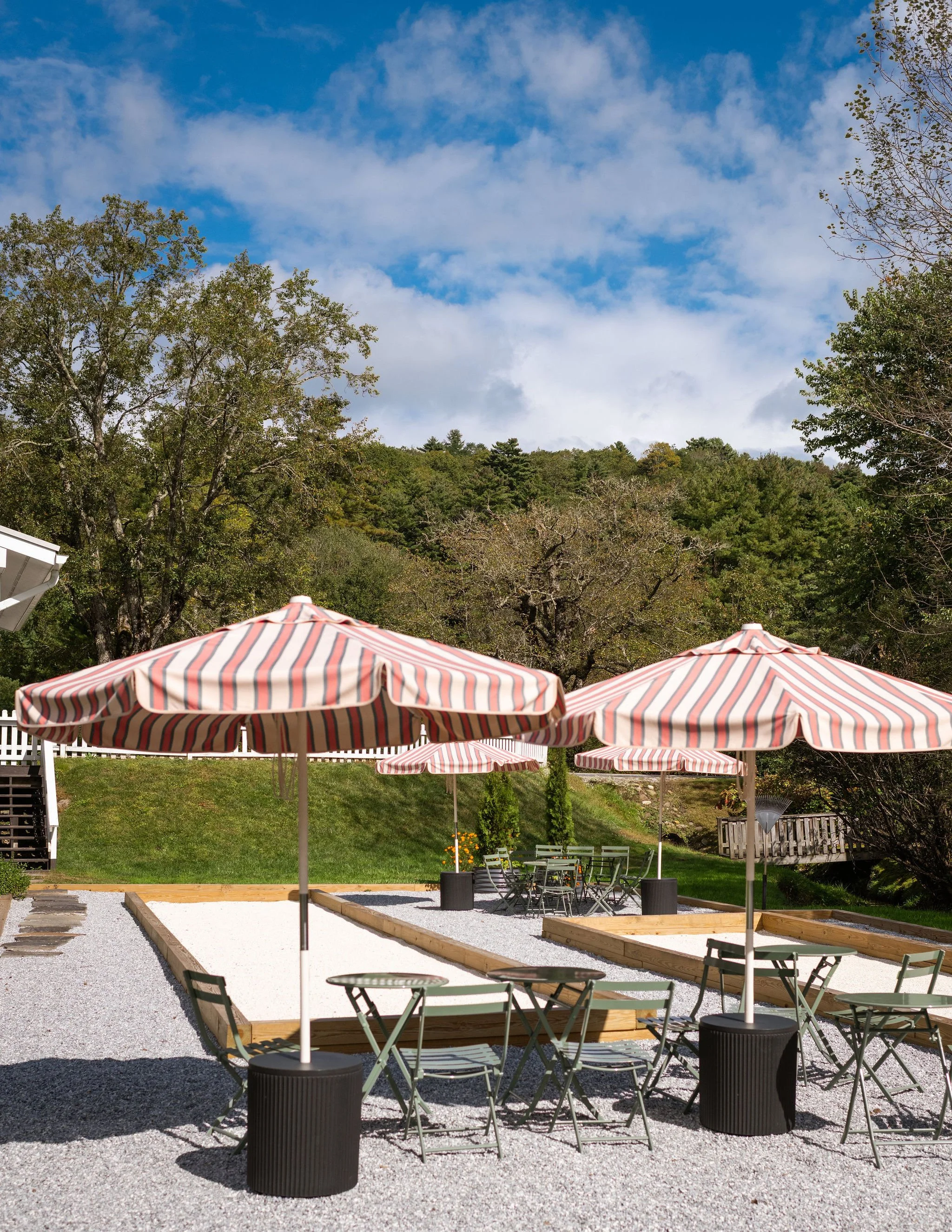 Outdoor patio with striped umbrellas, tables, and chairs on gravel, surrounded by greenery and trees under a partly cloudy sky.