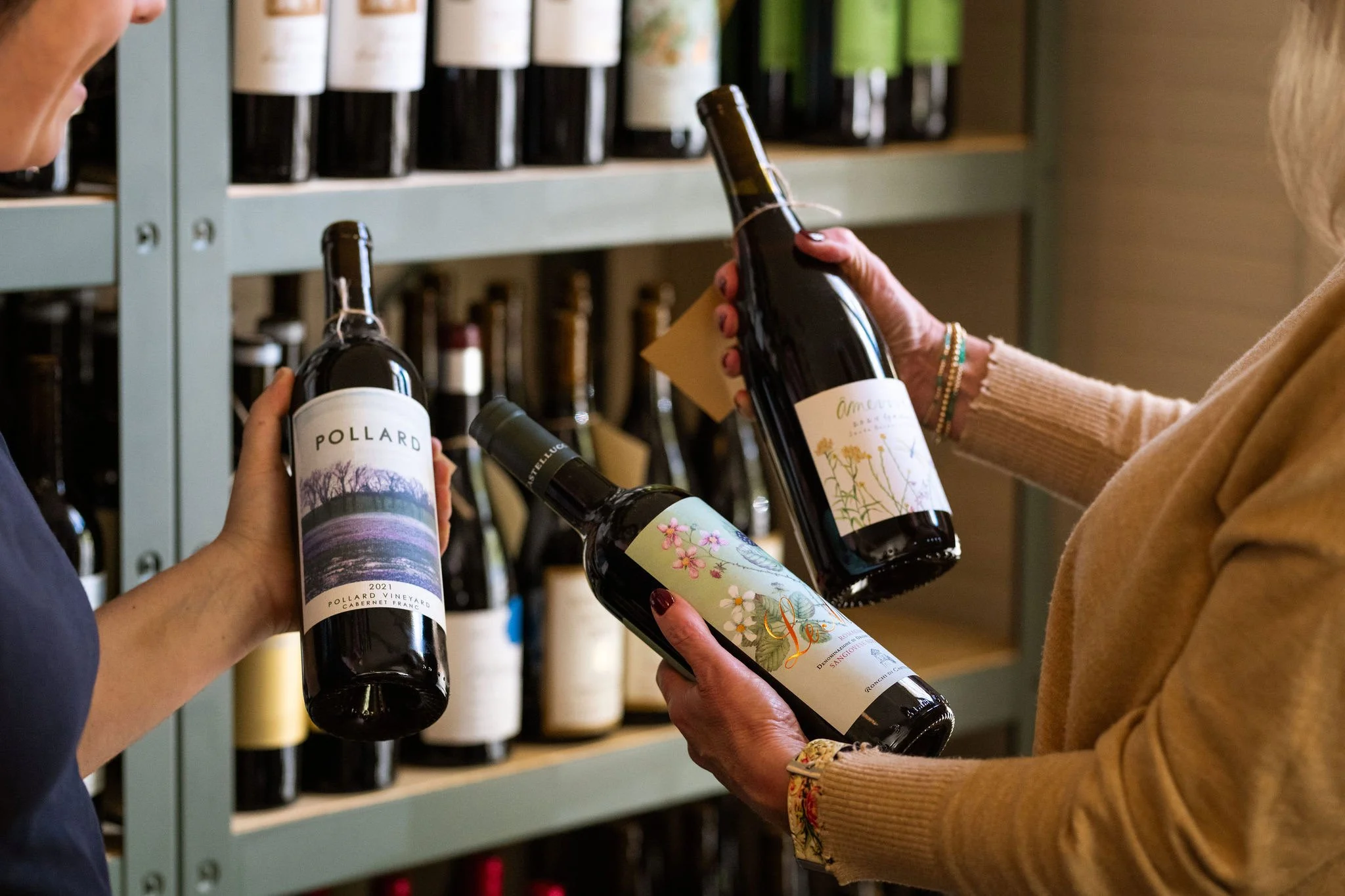 Two women are shopping for wine in a store. One woman holds a bottle with a landscape label, and the other woman is holding a bottle with a floral label. They are selecting wine from shelves filled with bottles.