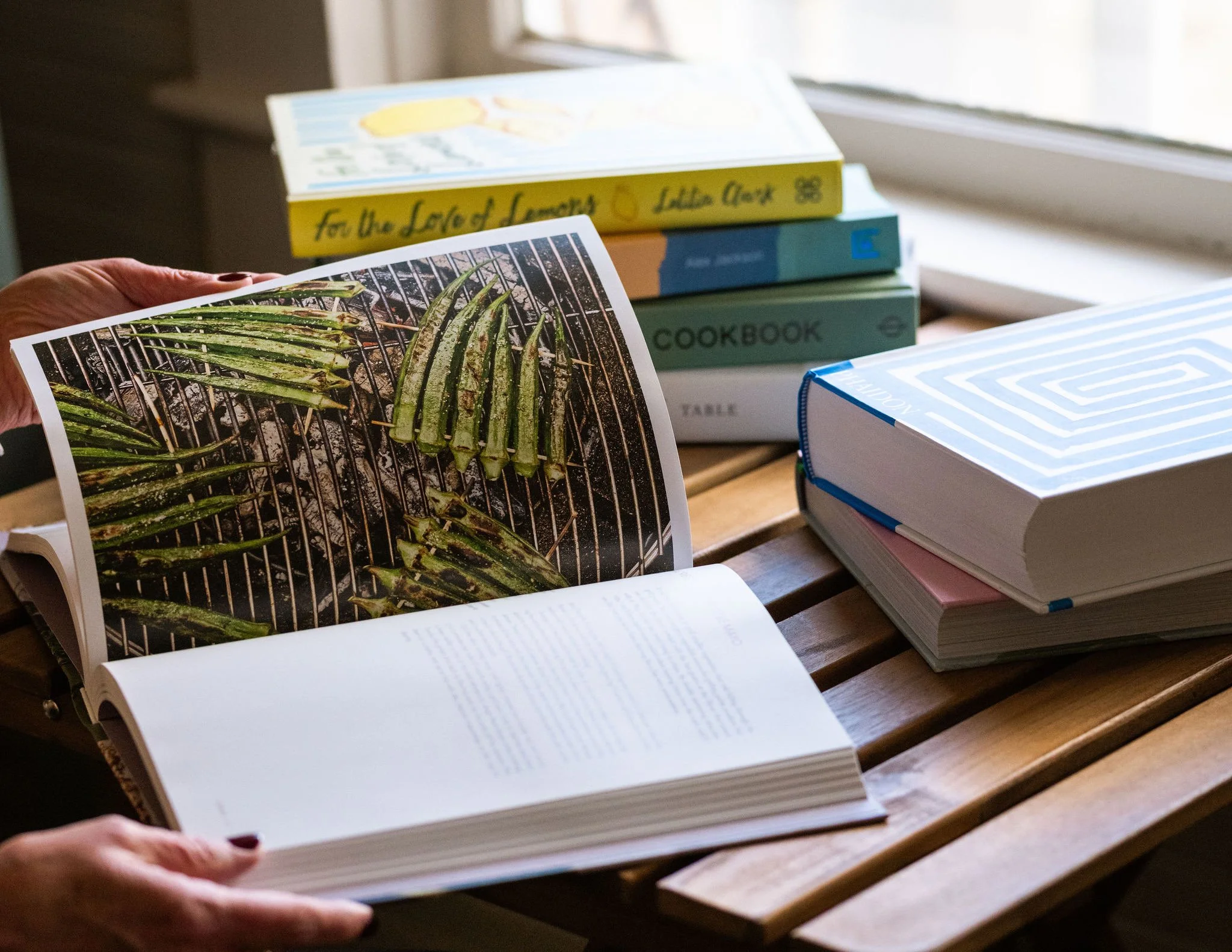 Open cookbook showing an image of grilled zucchini slices, with a stack of other books on a wooden table near a window.
