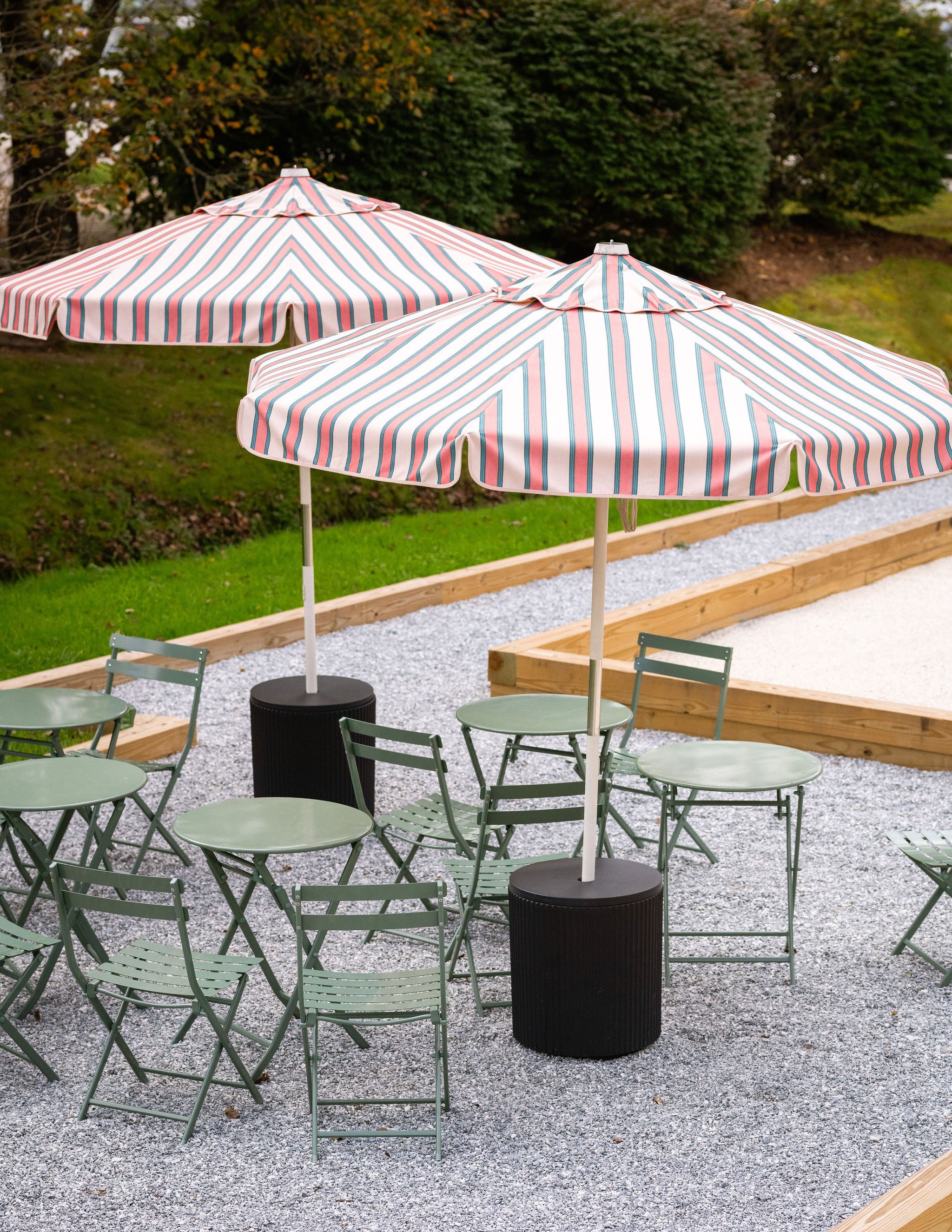 Outdoor seating area with green metal tables and chairs, two striped umbrellas, and a gravel ground. There is a wooden raised garden bed and trees in the background.