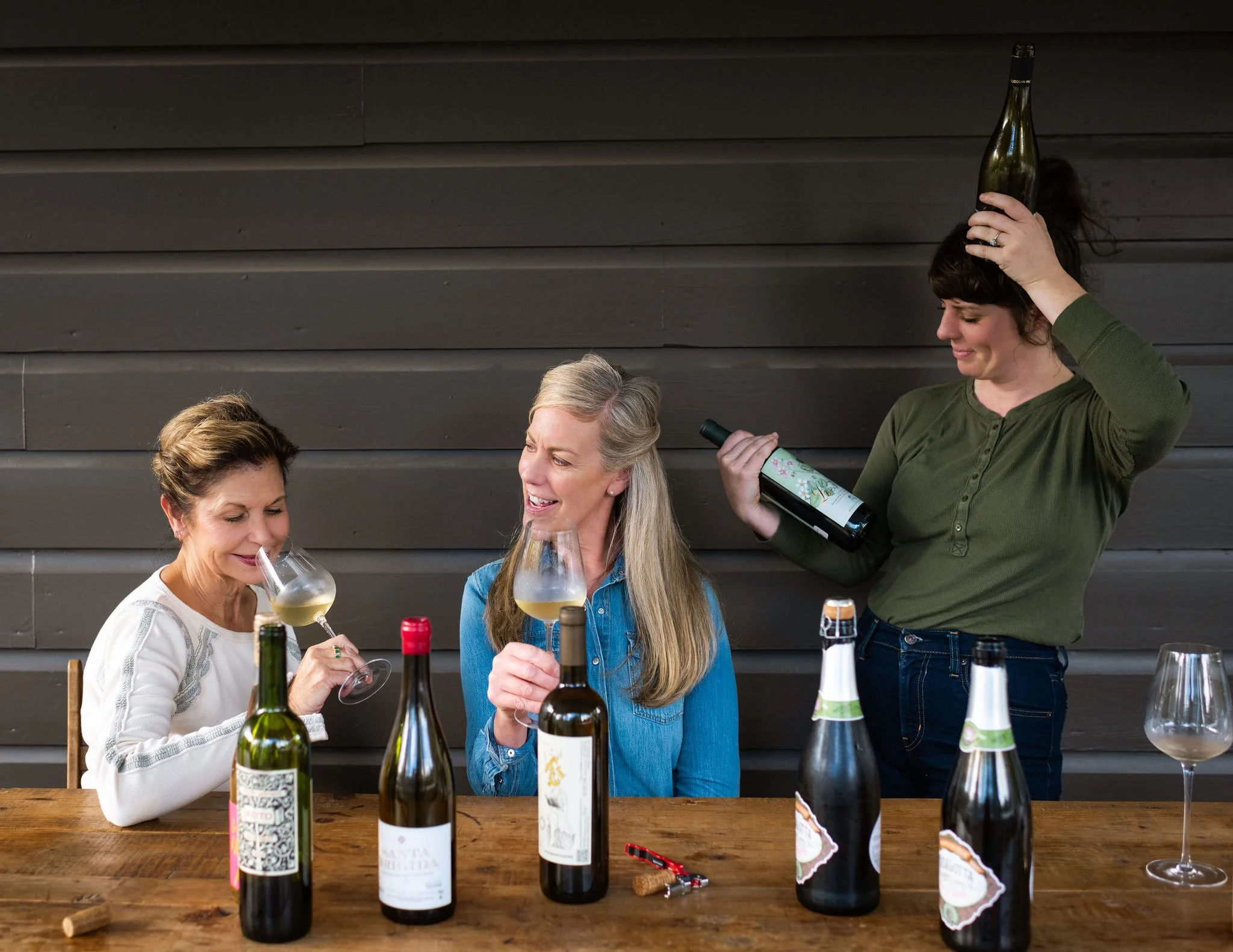 Three women enjoying wine at a wooden table with wine bottles, glasses, and wine openers, one woman balancing a wine bottle on her head.