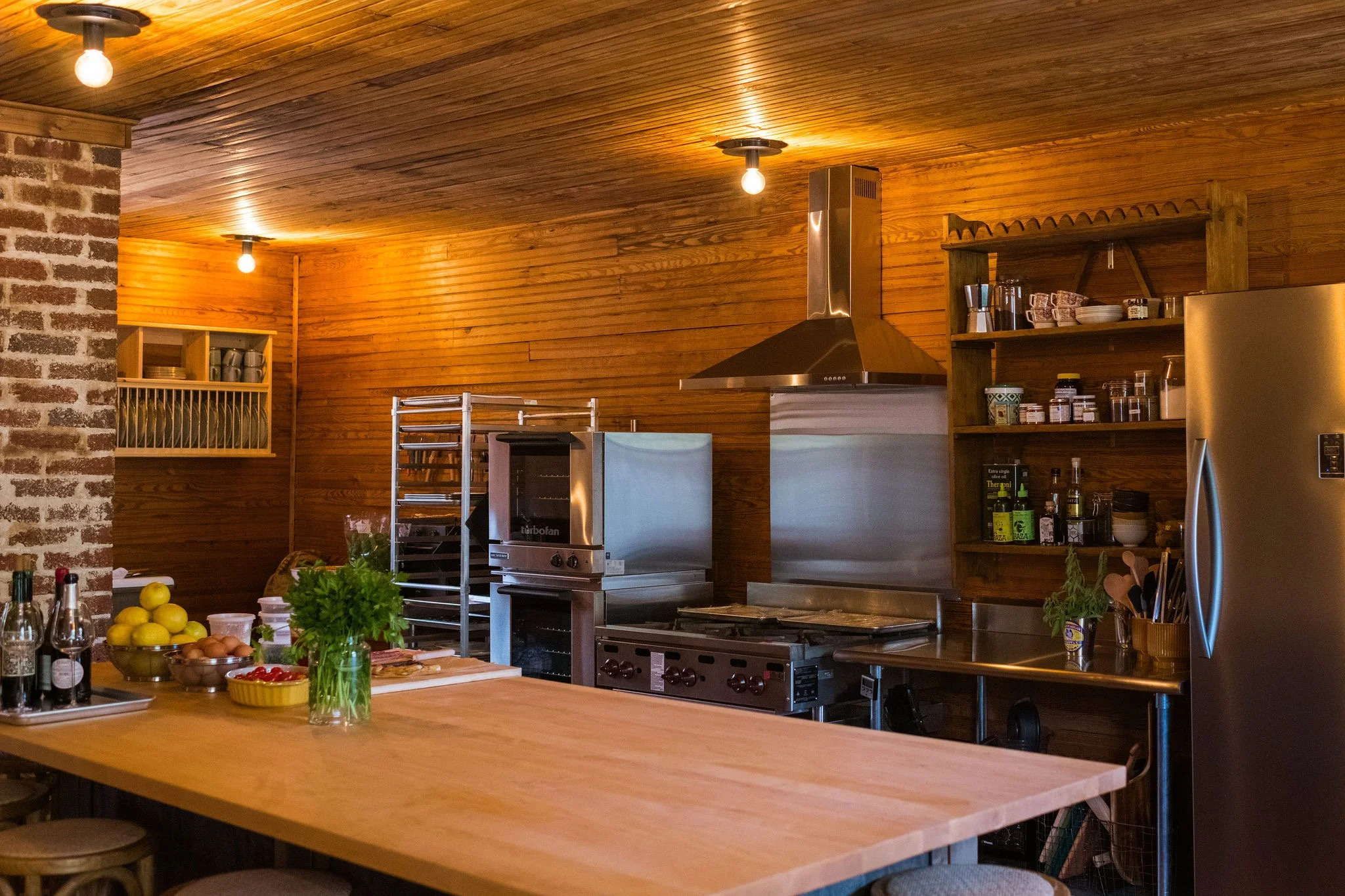 Kitchen with wooden walls and ceiling, stainless steel appliances, open shelves with dishes and spices, a large refrigerator, and a wooden island with seating.