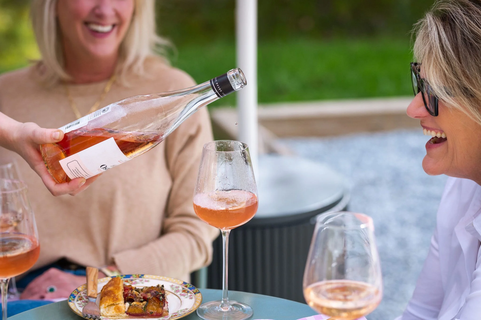 Two women celebrating with glasses of rosé wine at an outdoor gathering, one pouring wine from a bottle while the other smiles, with a plate of pizza on the table.