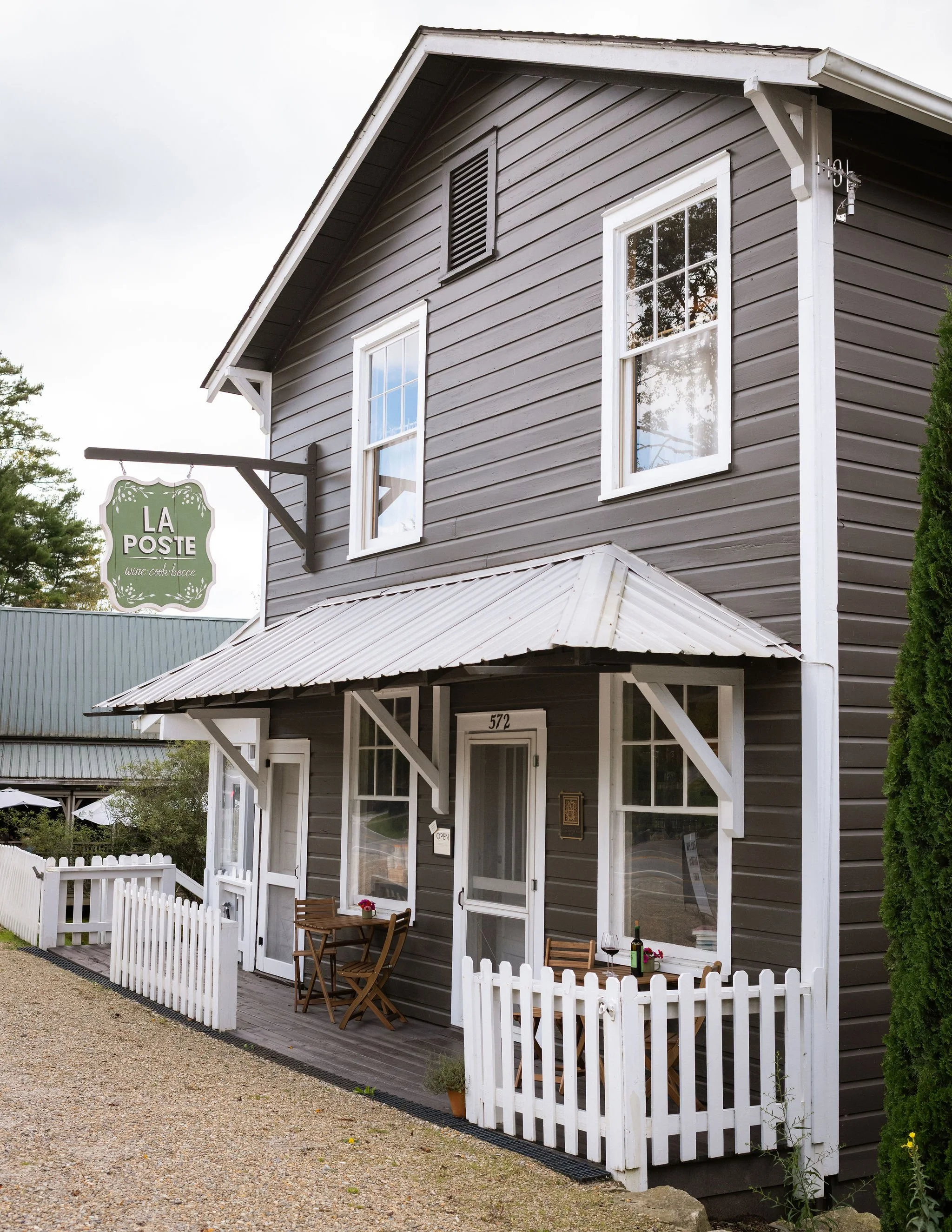 Gray two-story building with white trim and a small porch, outdoor table with chairs, bottle of wine, and wine glasses, white picket fence.