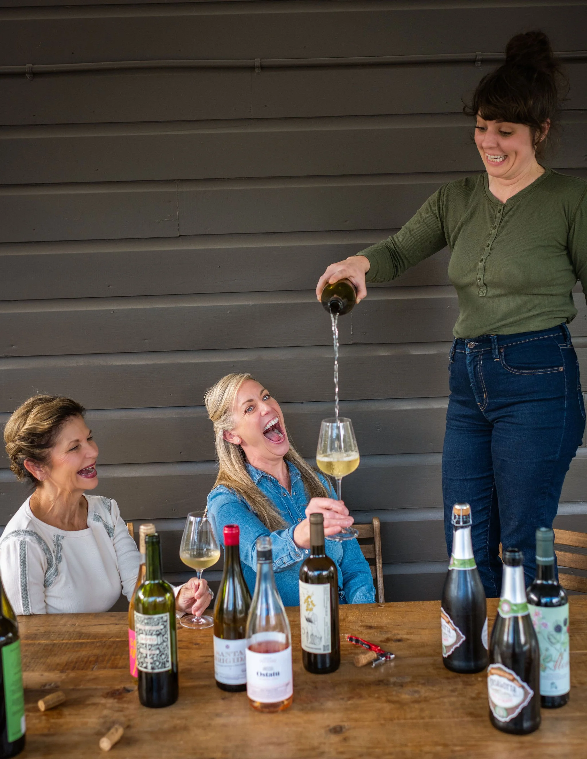 Three women enjoying a wine tasting, with one woman pouring white wine into another woman's glass while the third woman laughs, surrounded by various bottles of wine on the table.