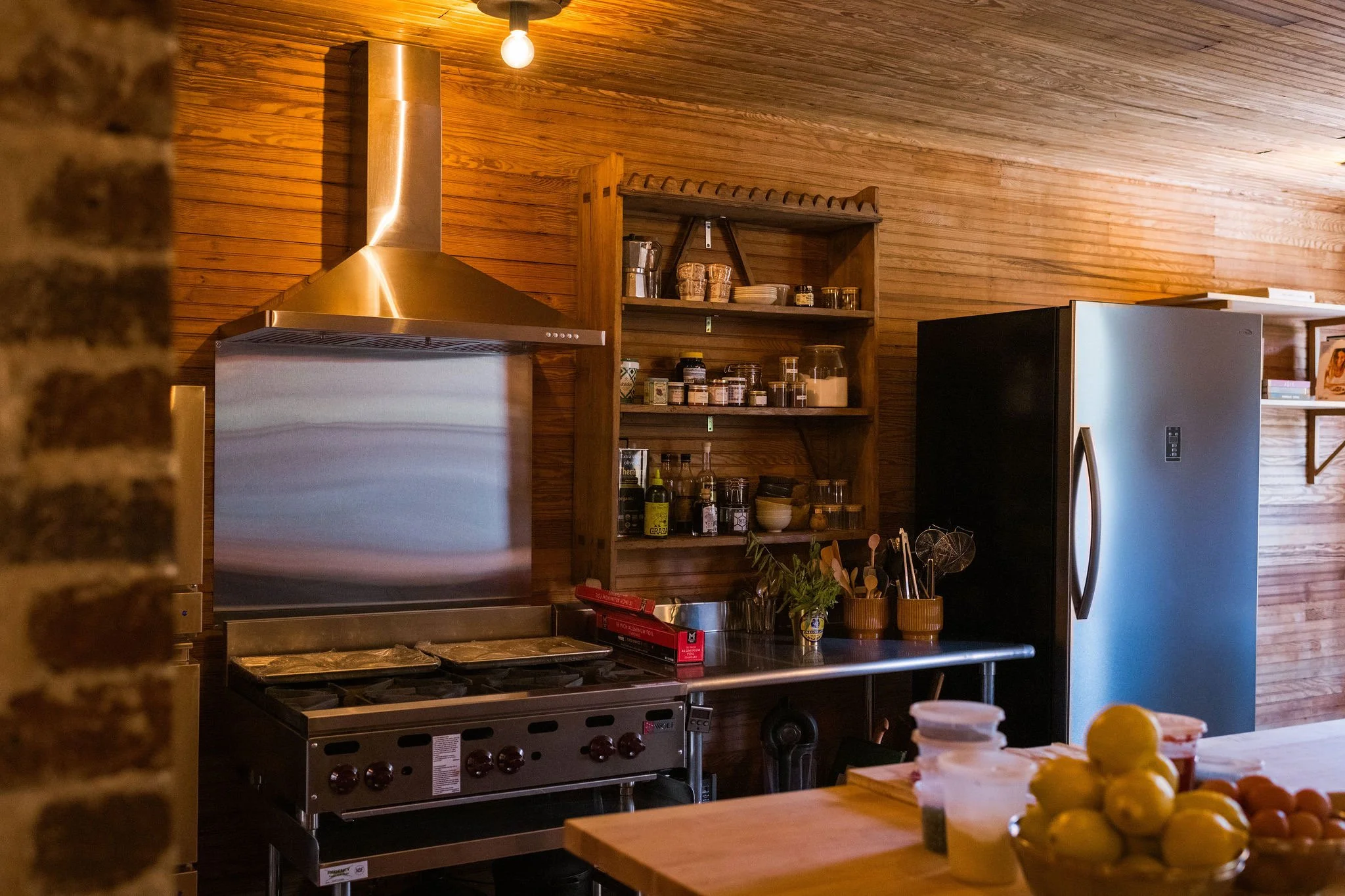 Kitchen with wooden walls, stainless steel range with hood, open shelving with jars, and a large silver refrigerator.