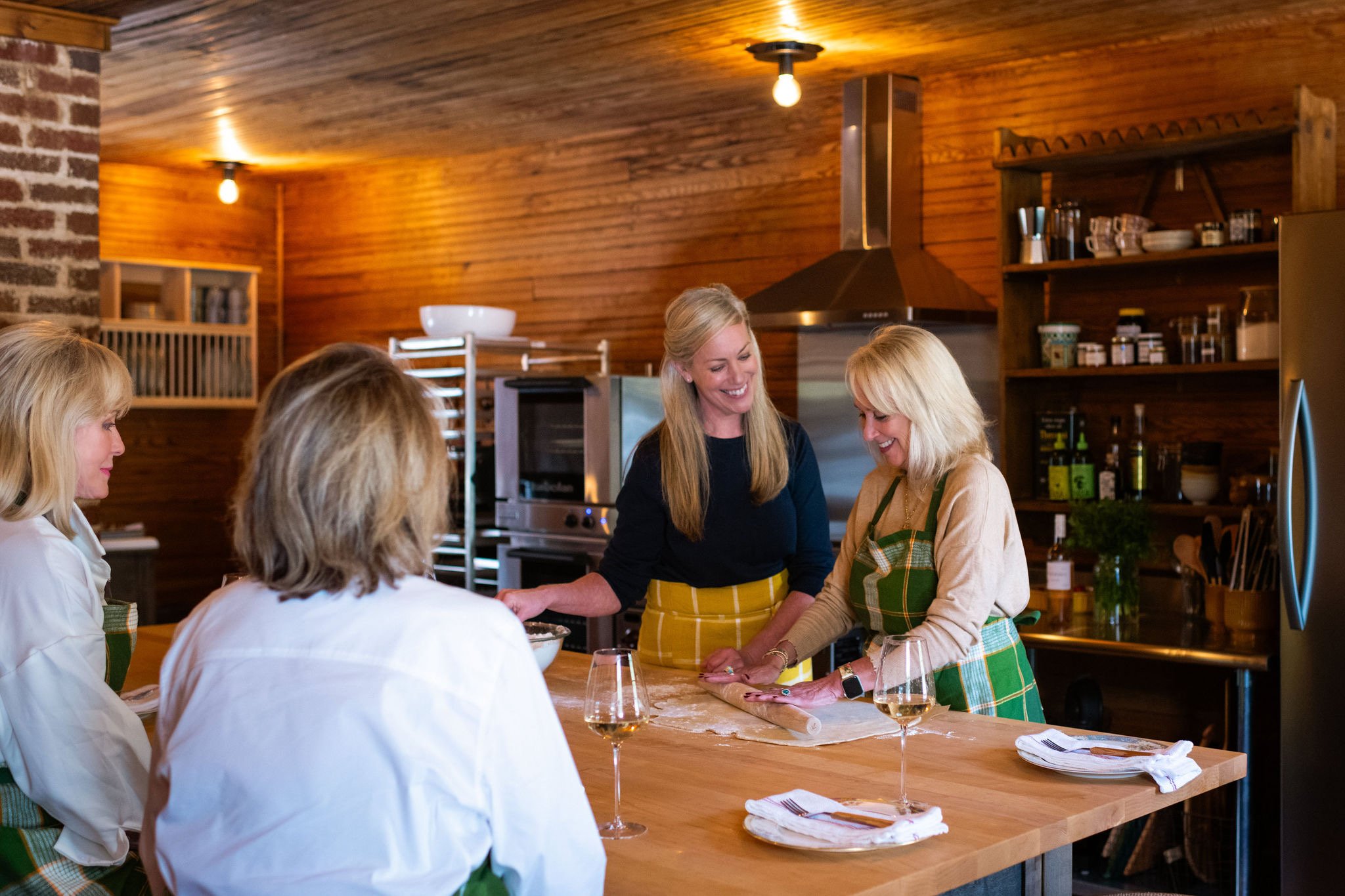 Four women in aprons gathered in a cozy, rustic kitchen with wooden walls, one rolling out dough while the others watch and smile.