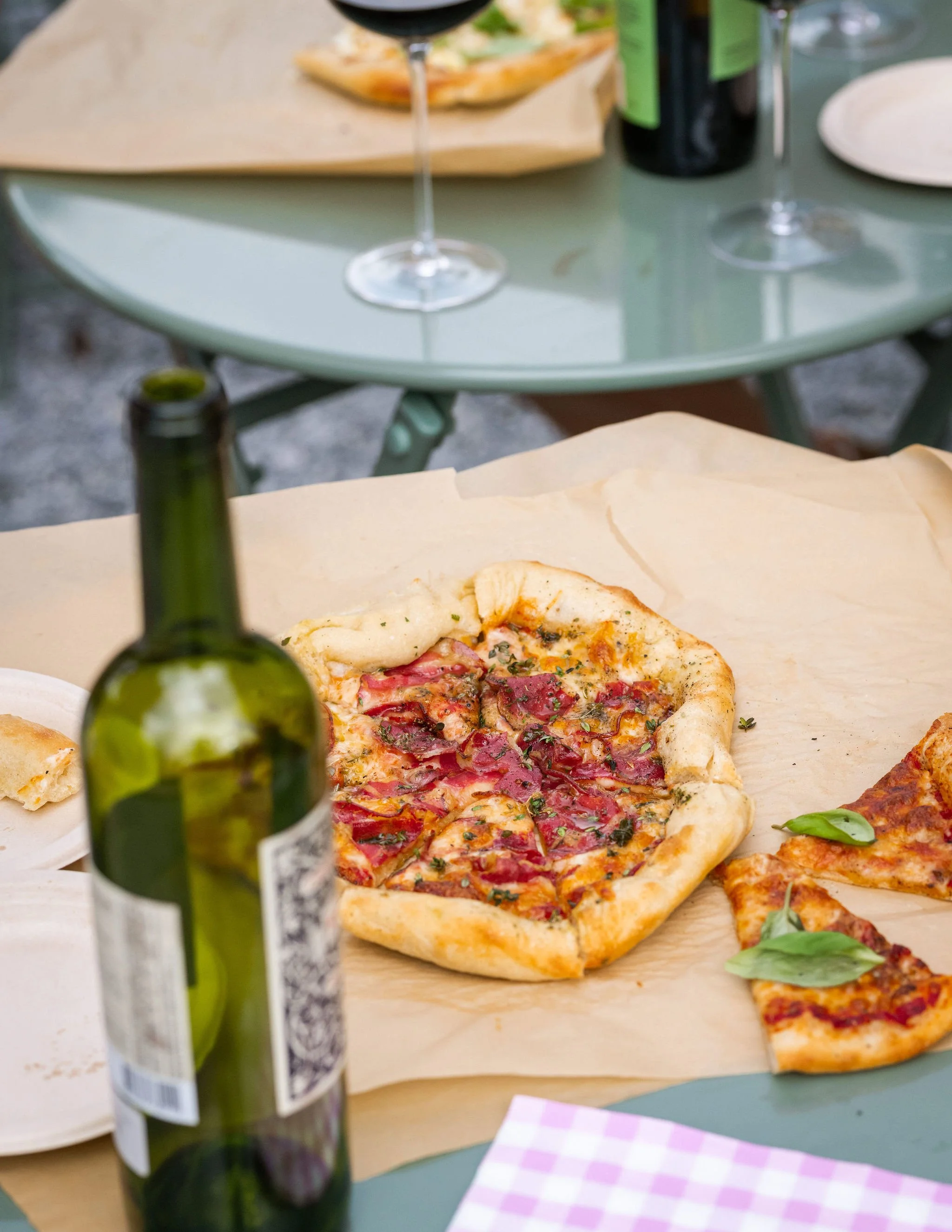Sliced pizza with pepperoni and basil on parchment paper, a wine bottle in the foreground, and a glass of red wine on a teal picnic table.