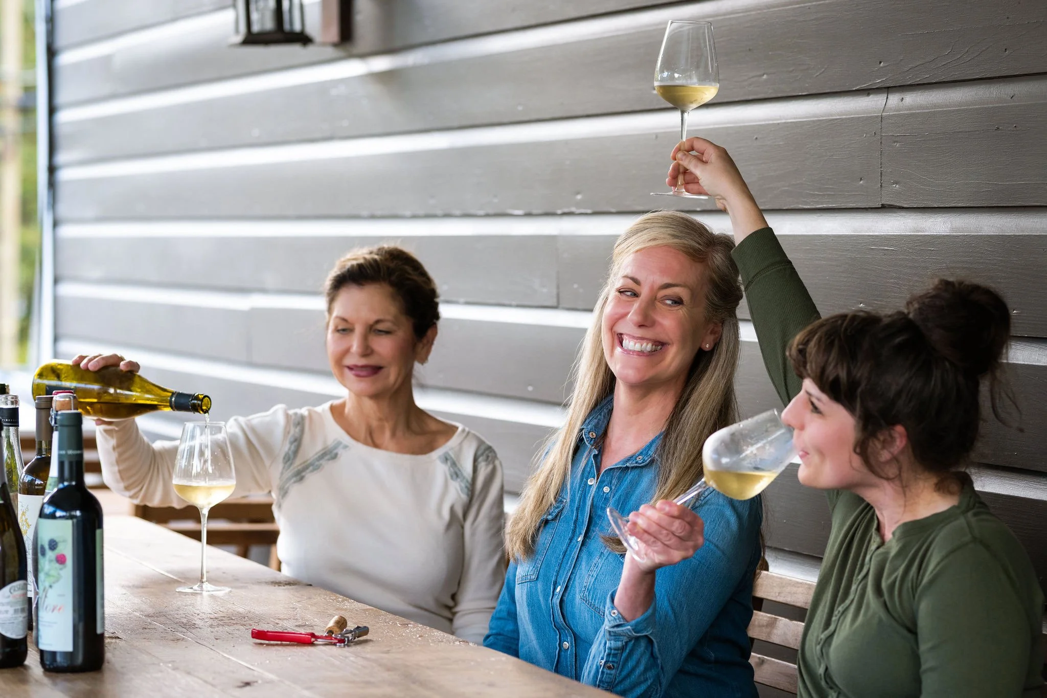 Three women sitting at a wooden table, enjoying glasses of white wine. The woman on the right is raising her glass and smiling, the woman in the middle is smiling and wearing a denim shirt, the woman on the left is pouring wine from a bottle. Bottles