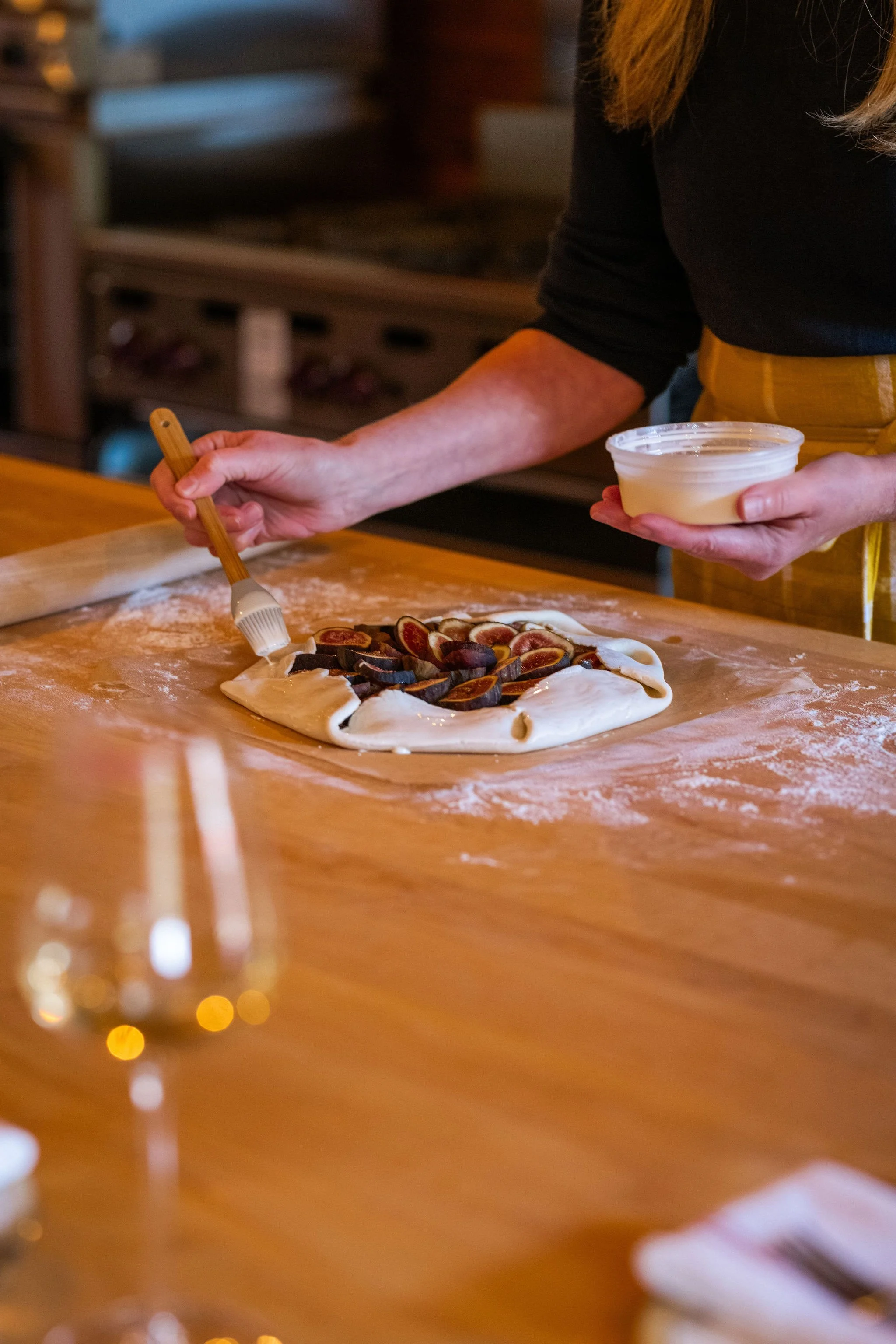 Person preparing a fig tart on a floured wooden countertop, brushing the dough with egg wash, holding a bowl of filling in one hand.