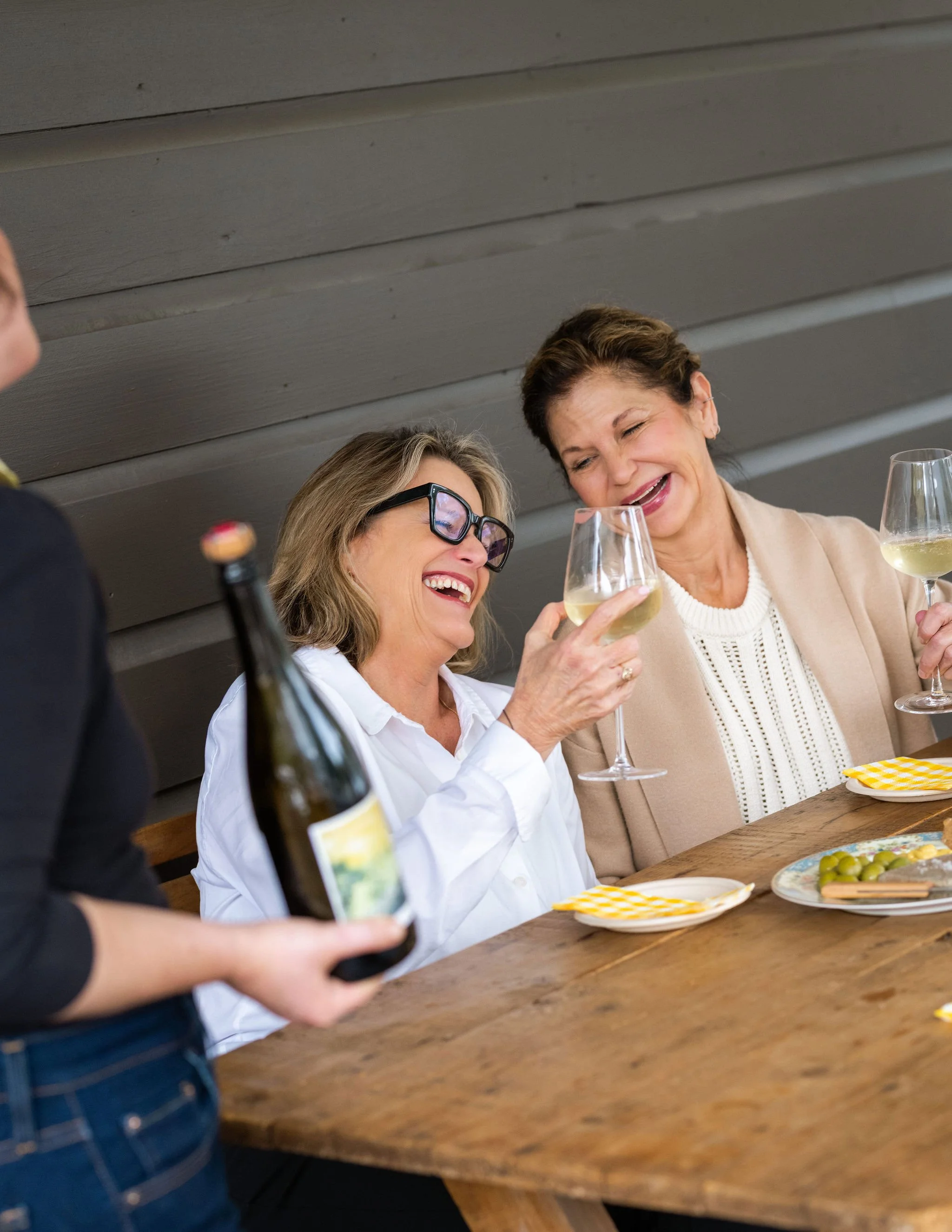 Two women smiling and raising wine glasses at a table with a server pouring wine