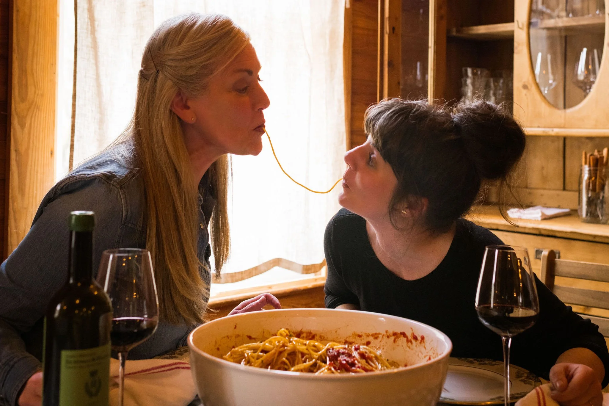 Two women at a dinner table with a bowl of pasta and glasses of red wine. One woman is pulling the other's face close with a strand of spaghetti between their lips.