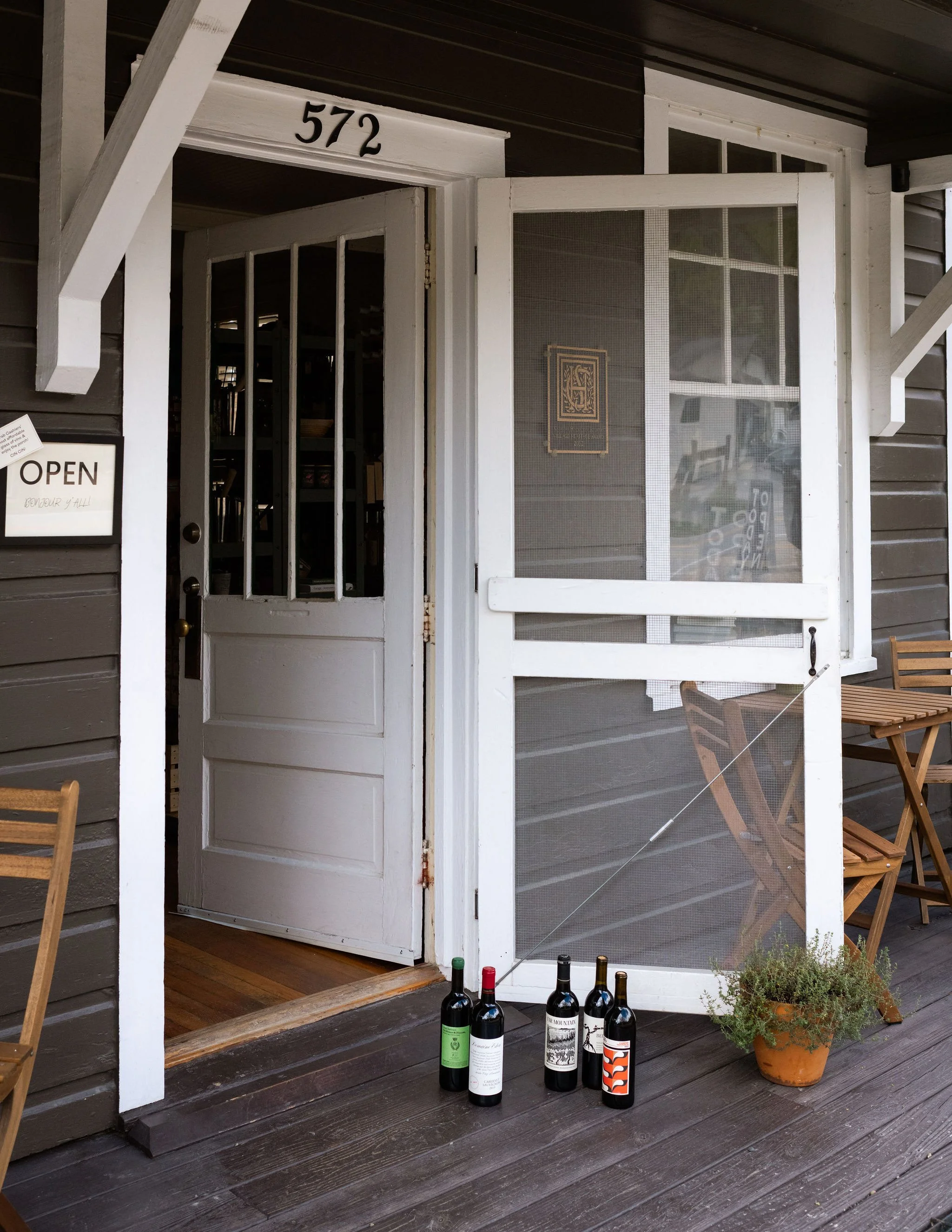 Front porch of a house with a white door, a screen door, and the house number 572. Seven bottles of wine, a potted plant, and two wooden chairs are on the porch.