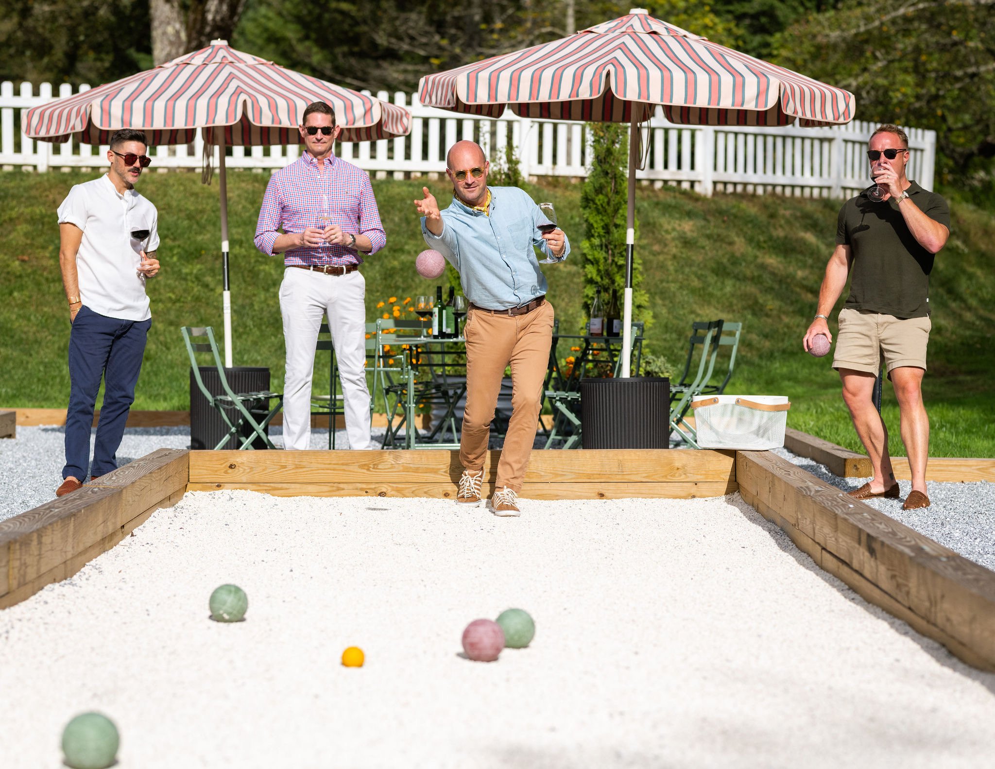 Four men playing bocce ball outdoors on a sunny day, with two on each side of a bocce court. Two are wearing sunglasses, and they are standing around the court with some holding drinks. Two umbrellas provide shade, and there is a table with bottles and glasses behind them.