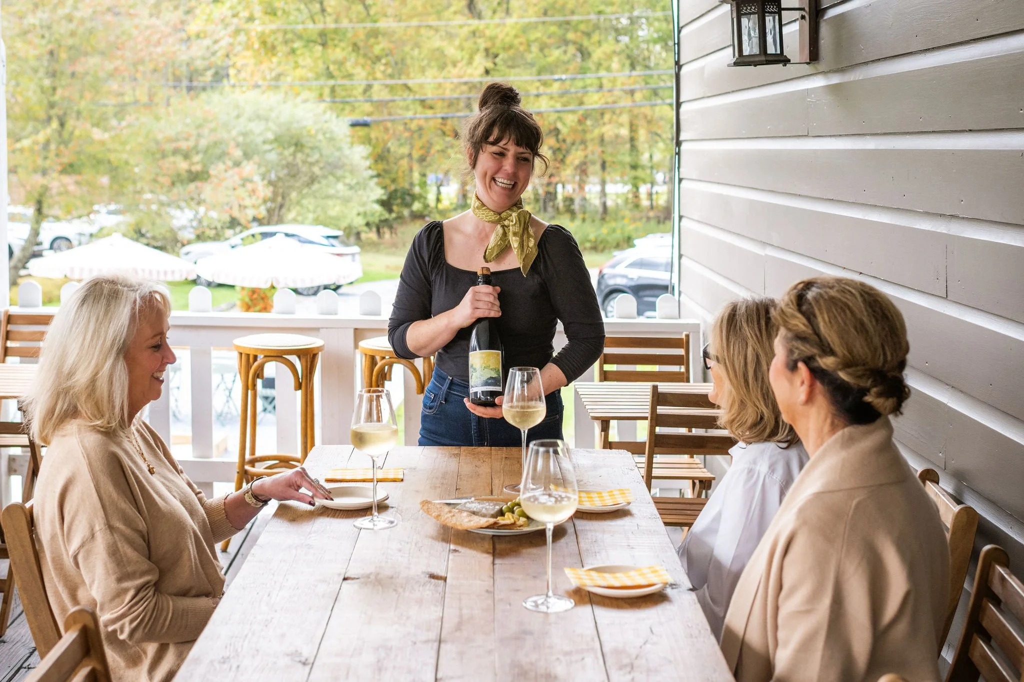 A woman serving wine to three women seated at a wooden table on a patio, with a scenic outdoor background.