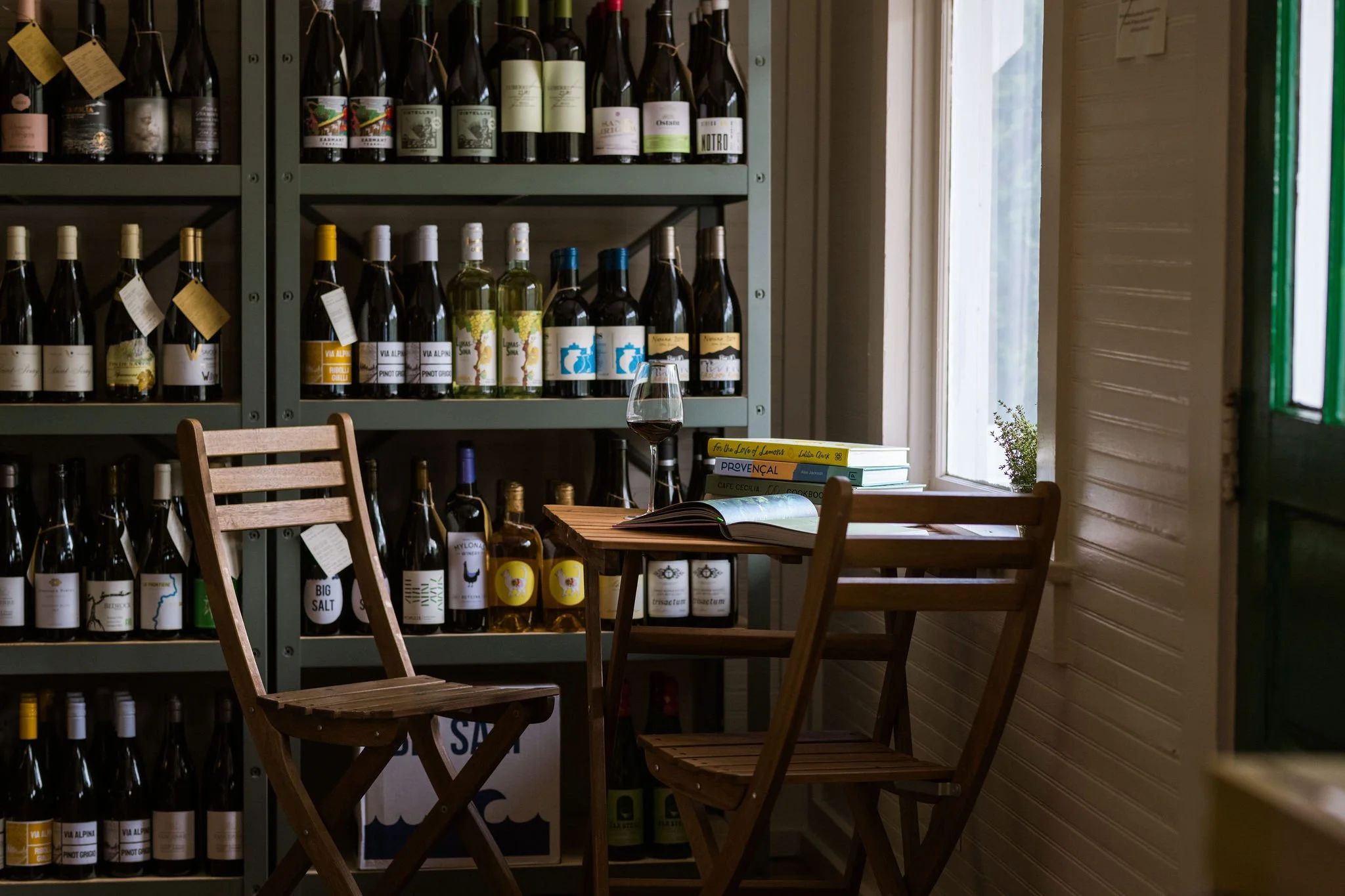 A small wooden table with two wooden chairs in a cozy corner of a wine shop or wine cellar. On the table, there is an open book, a glass of red wine, and a stack of books. Behind the table, there are shelves filled with various bottles of wine. A win