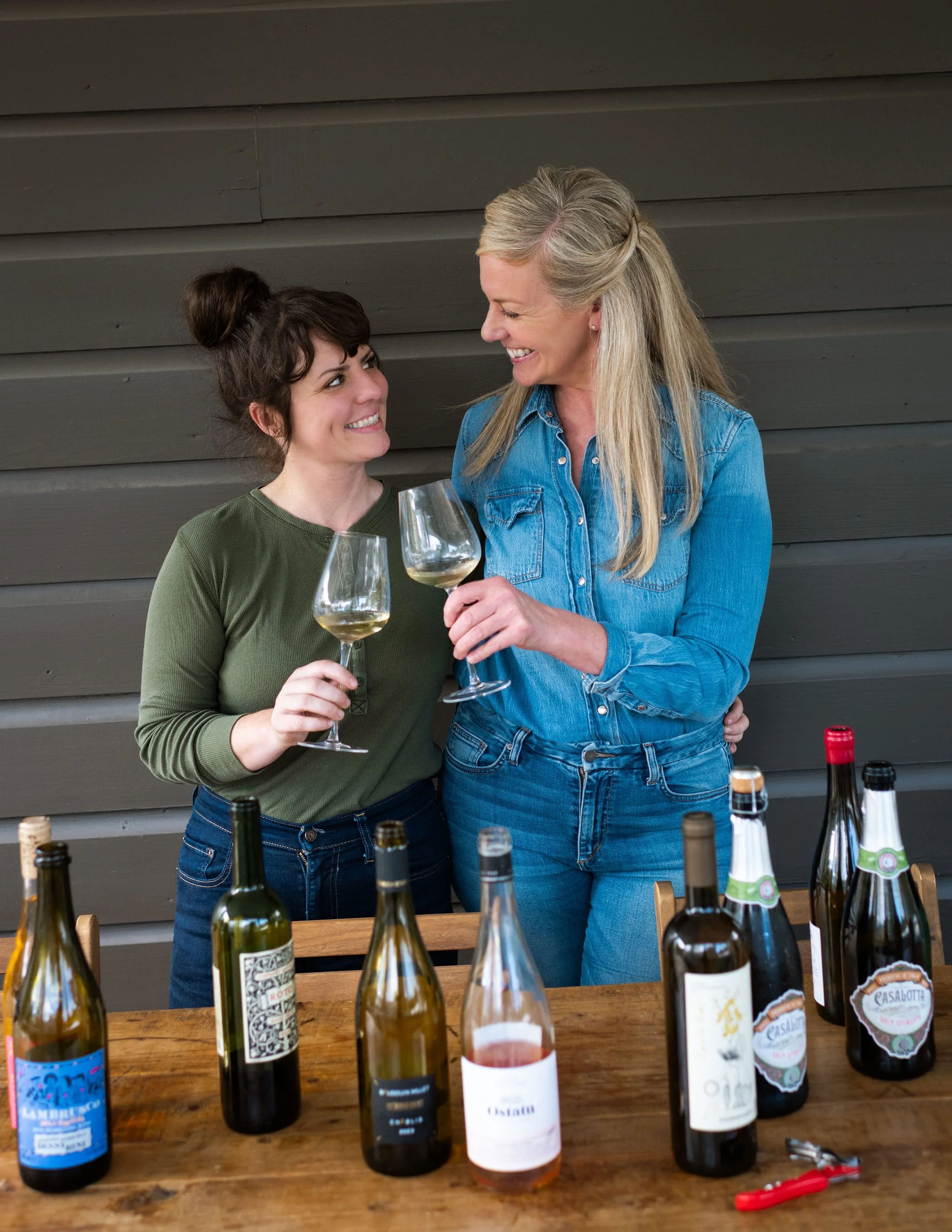 Two women enjoying wine together, holding glasses, with bottles of wine on a table in front of them.