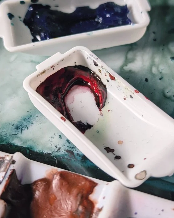 A photograph of two watercolour pans resting on the mixing area of my main watercolour palette. The pan in the background contains Phthalo Blue green shade, and the pan to the front contains Perylene Dark Red.