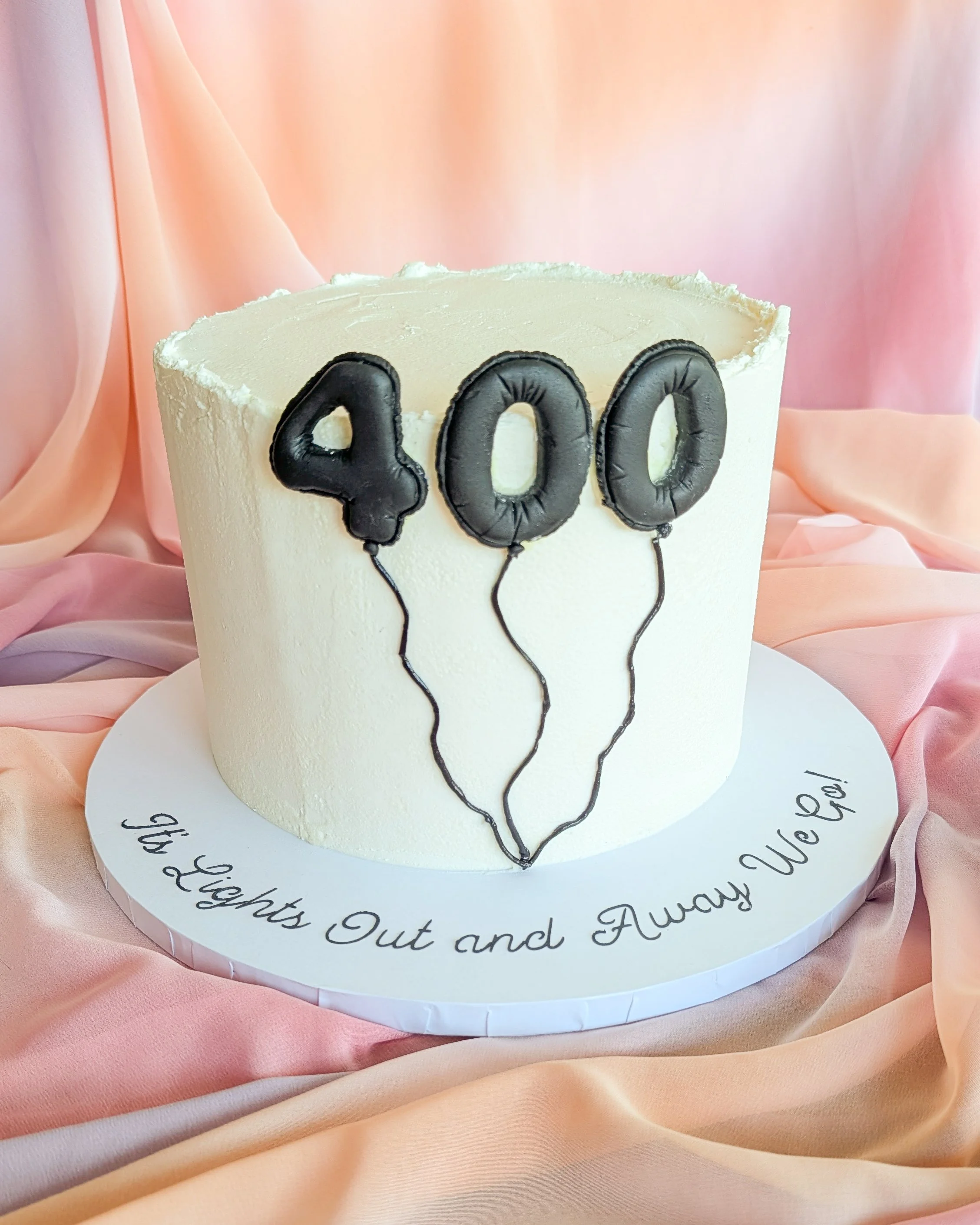 White cake with black number balloons spelling '400', decorated with black strings, placed on a white cake board with cursive writing, surrounded by peach-colored satin fabric.
