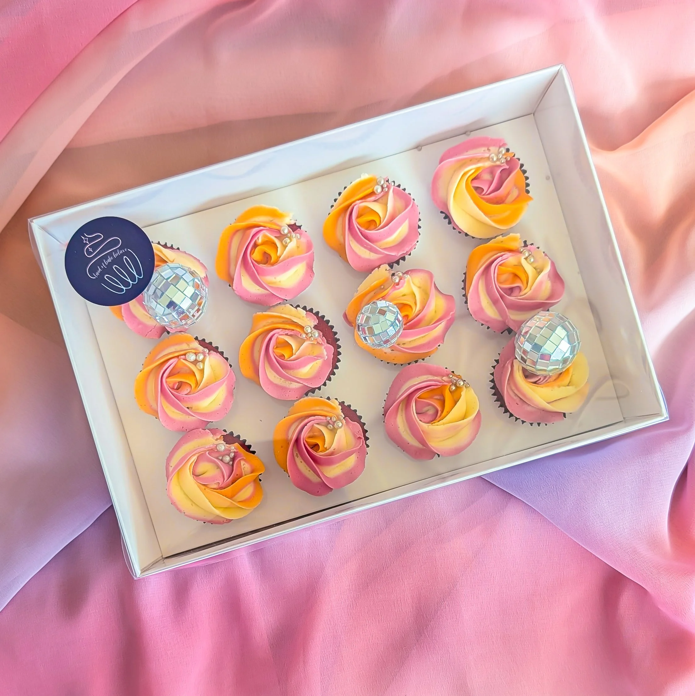 Box of cupcakes decorated with pink and yellow swirl frosting, adorned with small pearl-like and disco ball decorations, on a pink fabric surface.