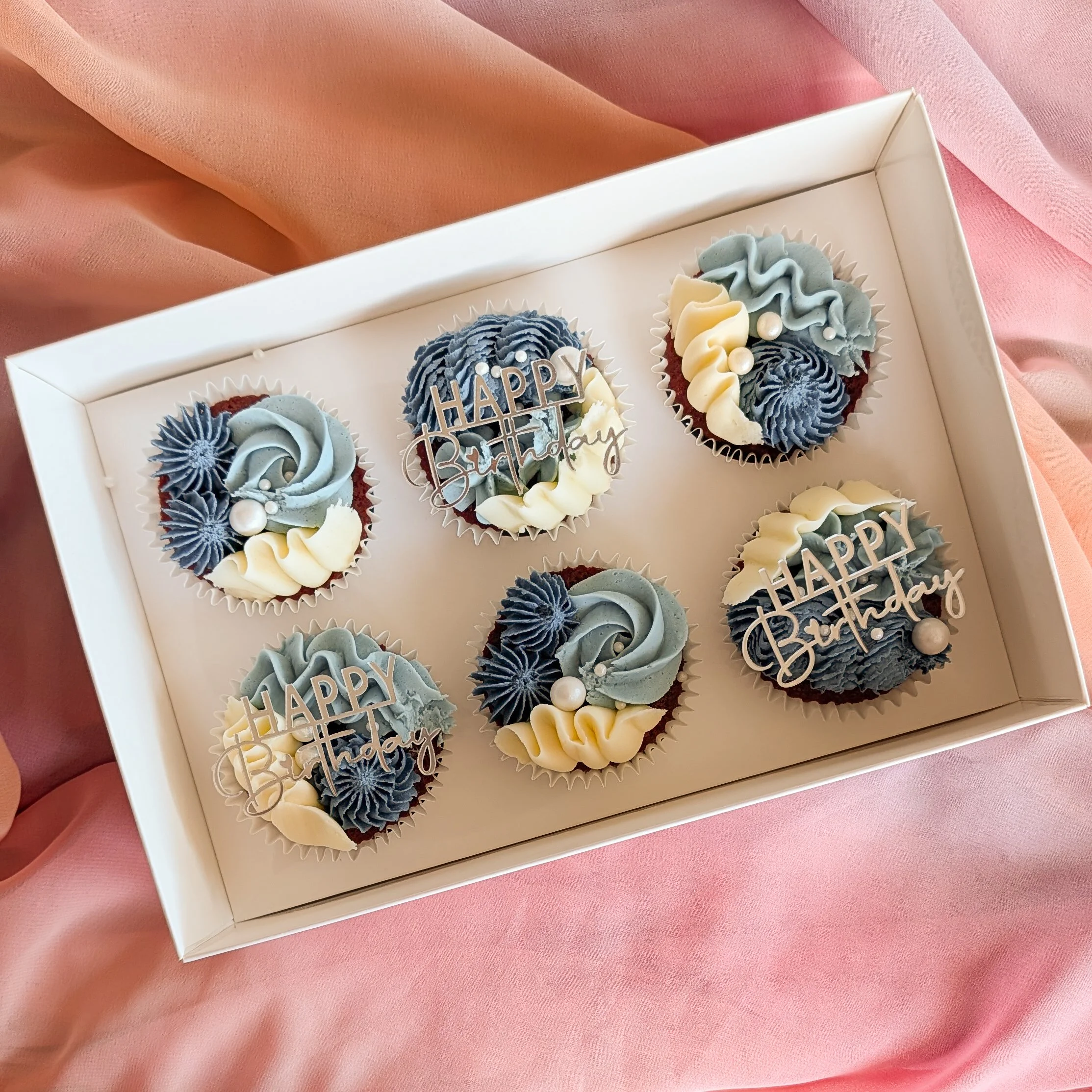 A white box containing six decorated cupcakes with blue and white frosting, pearls, and birthday message toppers on a pink fabric surface.