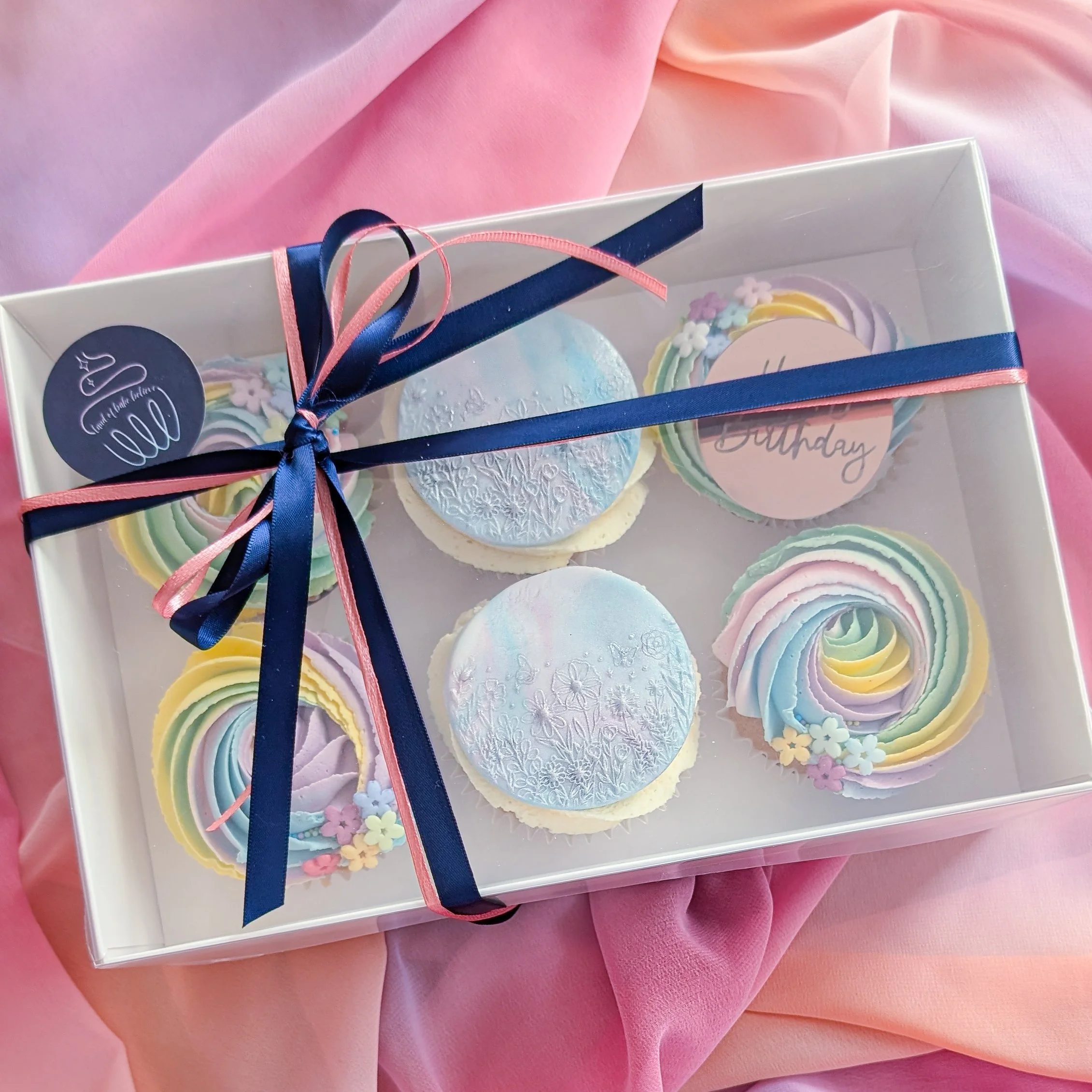 A rectangular box with colorful Christmas cookies decorated with pastel icing, tied with pink and navy blue ribbons, sitting on pink fabric. The box has a round label that says 'Sweet Glitter.'