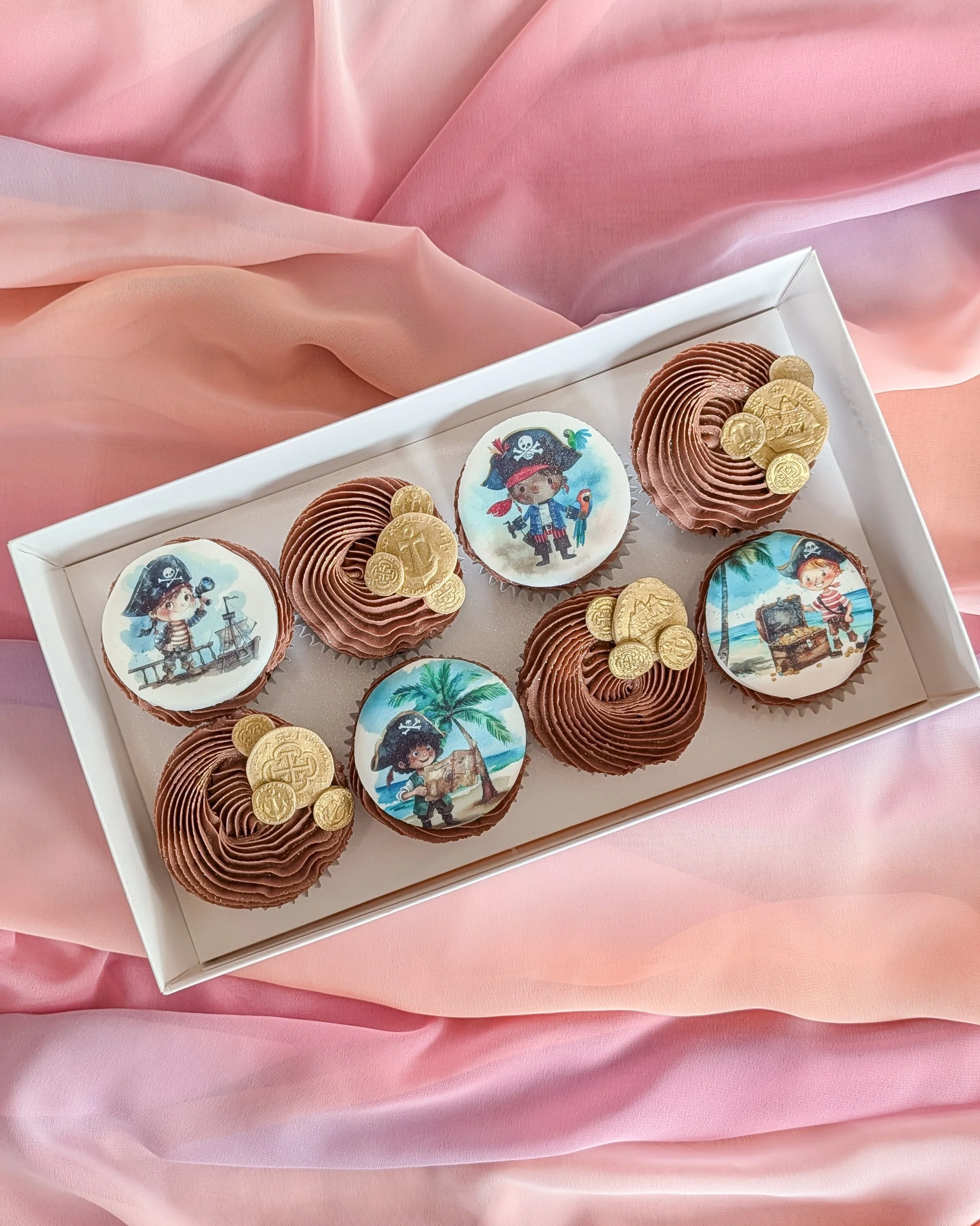 Box of chocolates decorated with pirate-themed edible toppers and gold coins on pink fabric background.