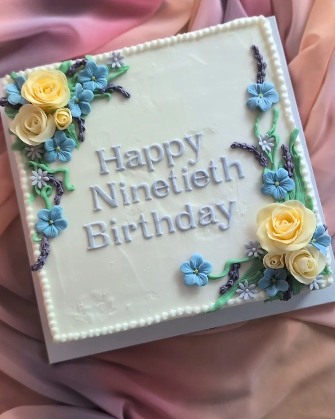 A rectangular birthday cake with white frosting decorated with yellow and blue flowers and green leaves. The cake has the message 'Happy Ninetieth Birthday' written in white icing.