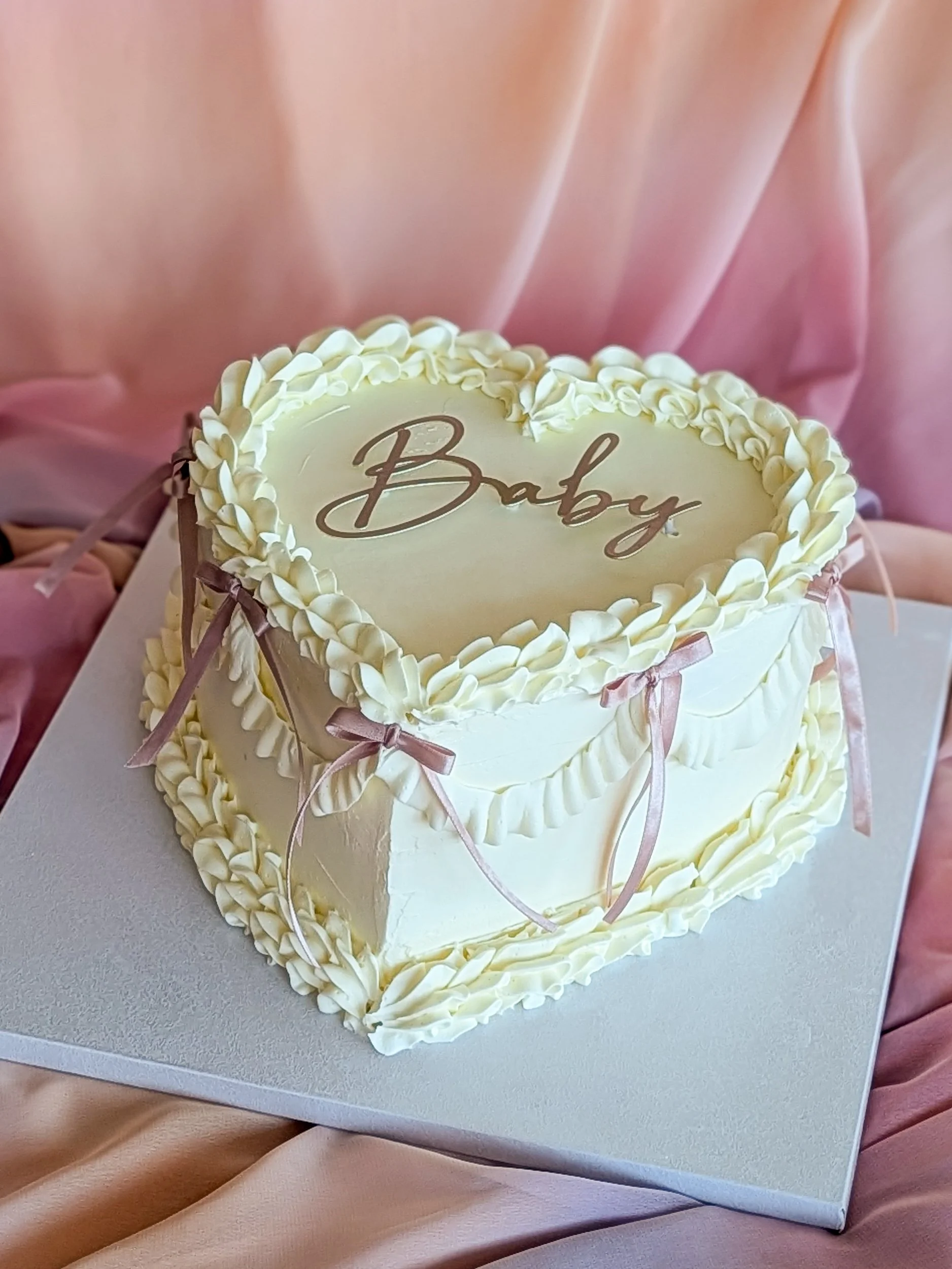 Heart-shaped birthday cake with white frosting and ribbon decorations, with the word 'Baby' written on top, being held by a person in pink clothing.