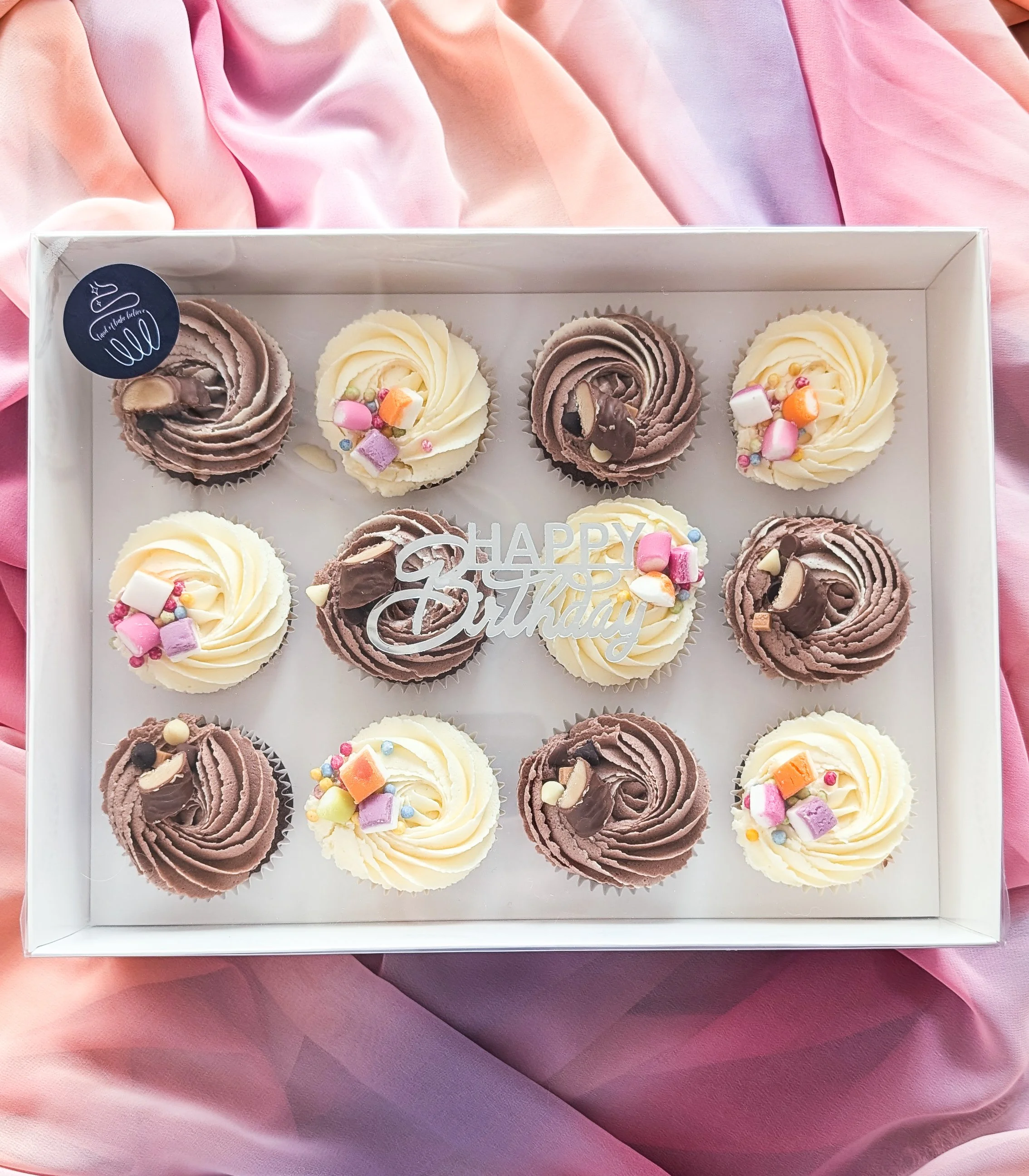 Box of assorted cupcakes with pink, white, and brown frosting, topped with colorful sprinkles and candies, with a 'Happy Birthday' decoration inside the box, on a pink fabric background.