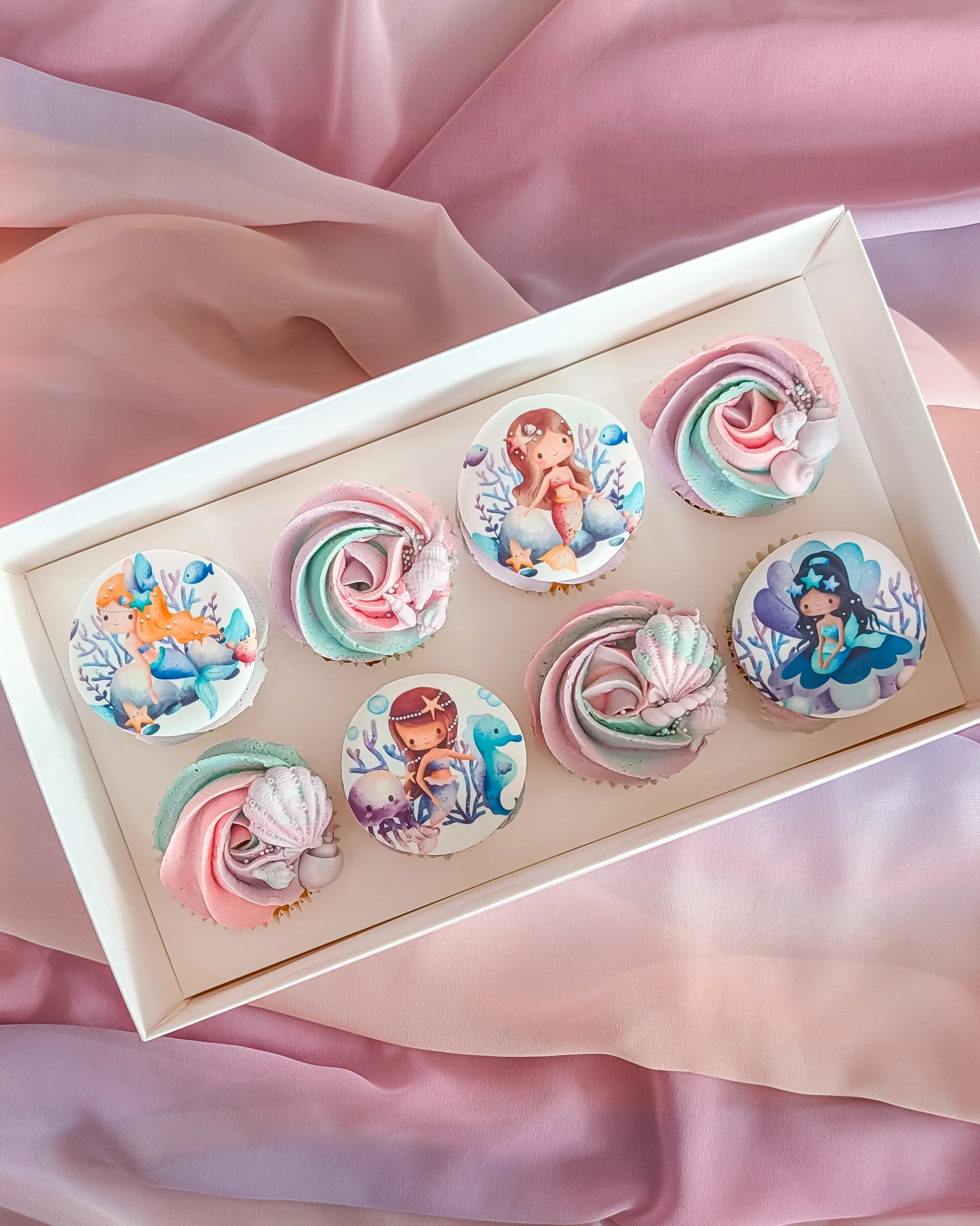 A white tray of cupcakes with mermaid-themed and ocean-themed edible images on top, surrounded by pink and purple swirled frosting, resting on pink fabric.