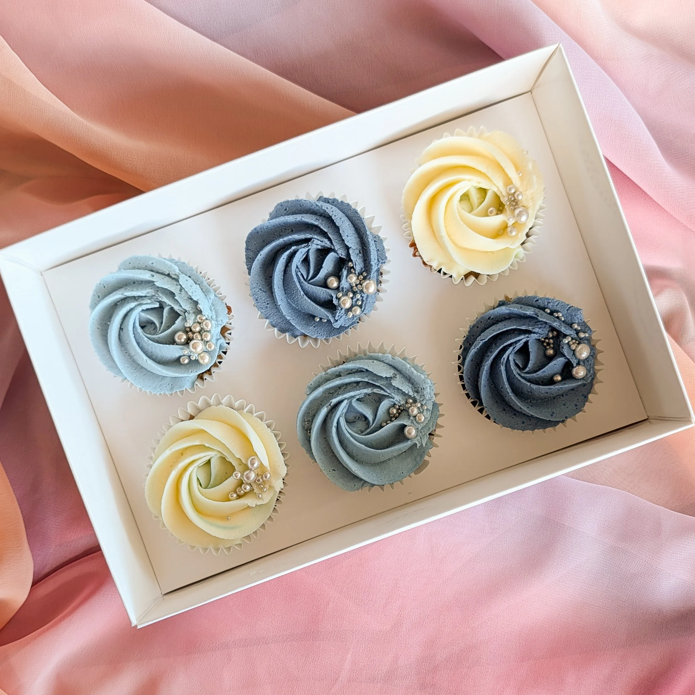 A white box of cupcakes decorated with swirls of purple and yellow frosting, each topped with edible silver pearls, on a pink fabric background.