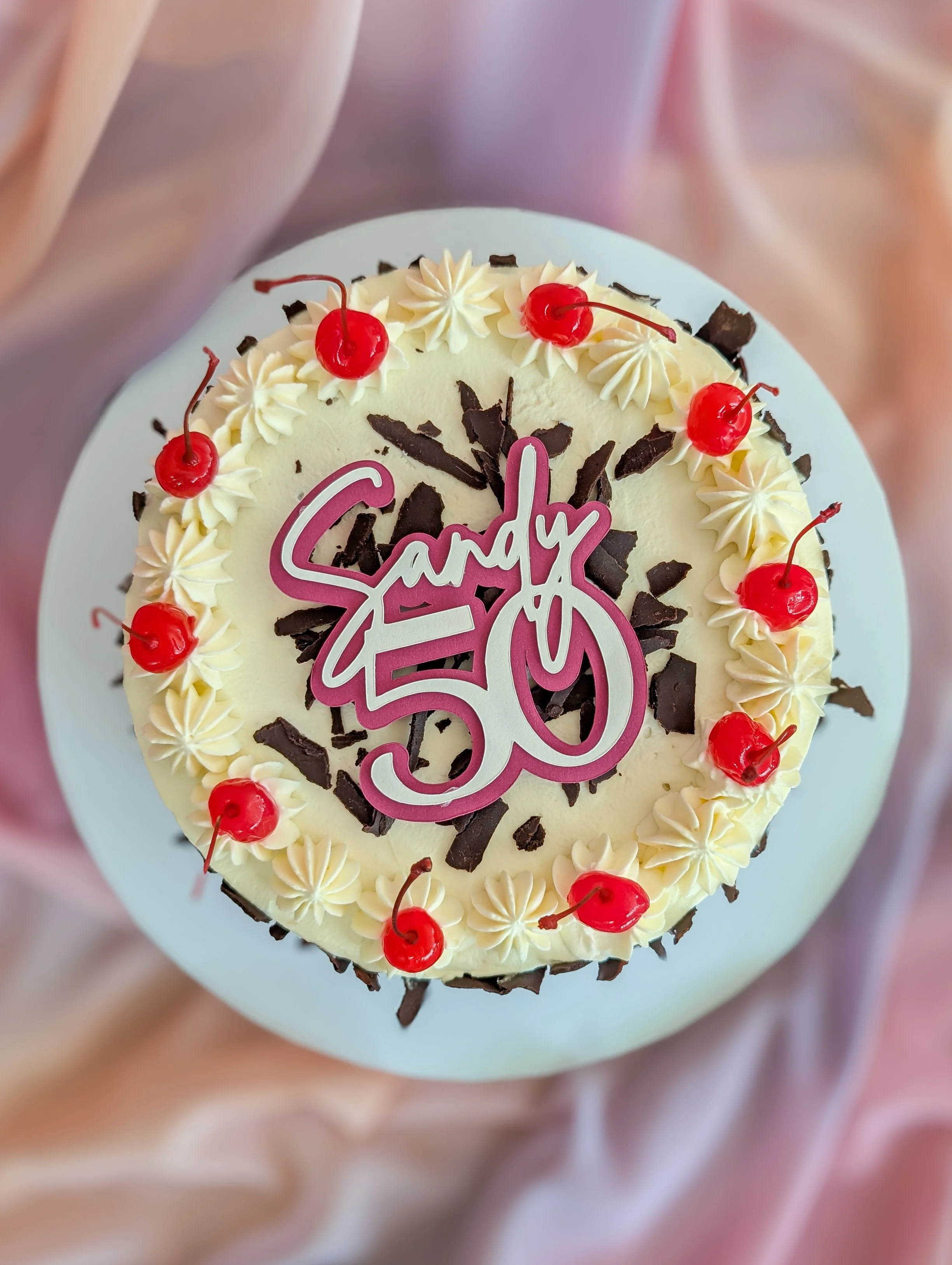 A birthday cake decorated with cherries and chocolate shavings, with a pink and white number 50 candle on top.
