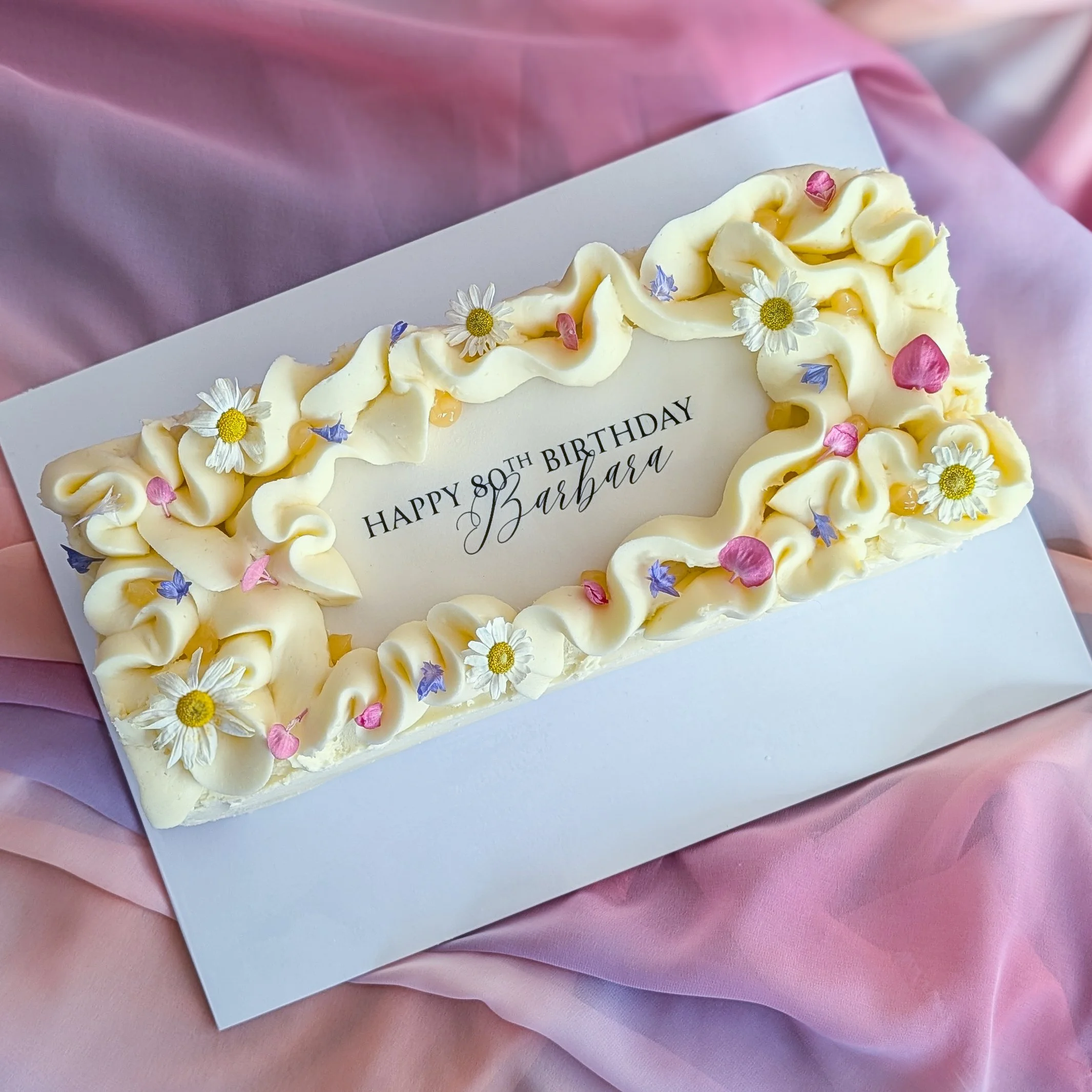 A birthday cake decorated with white frosting, edible flowers, pink and purple petals, and a message celebrating Barbara's 80th birthday.