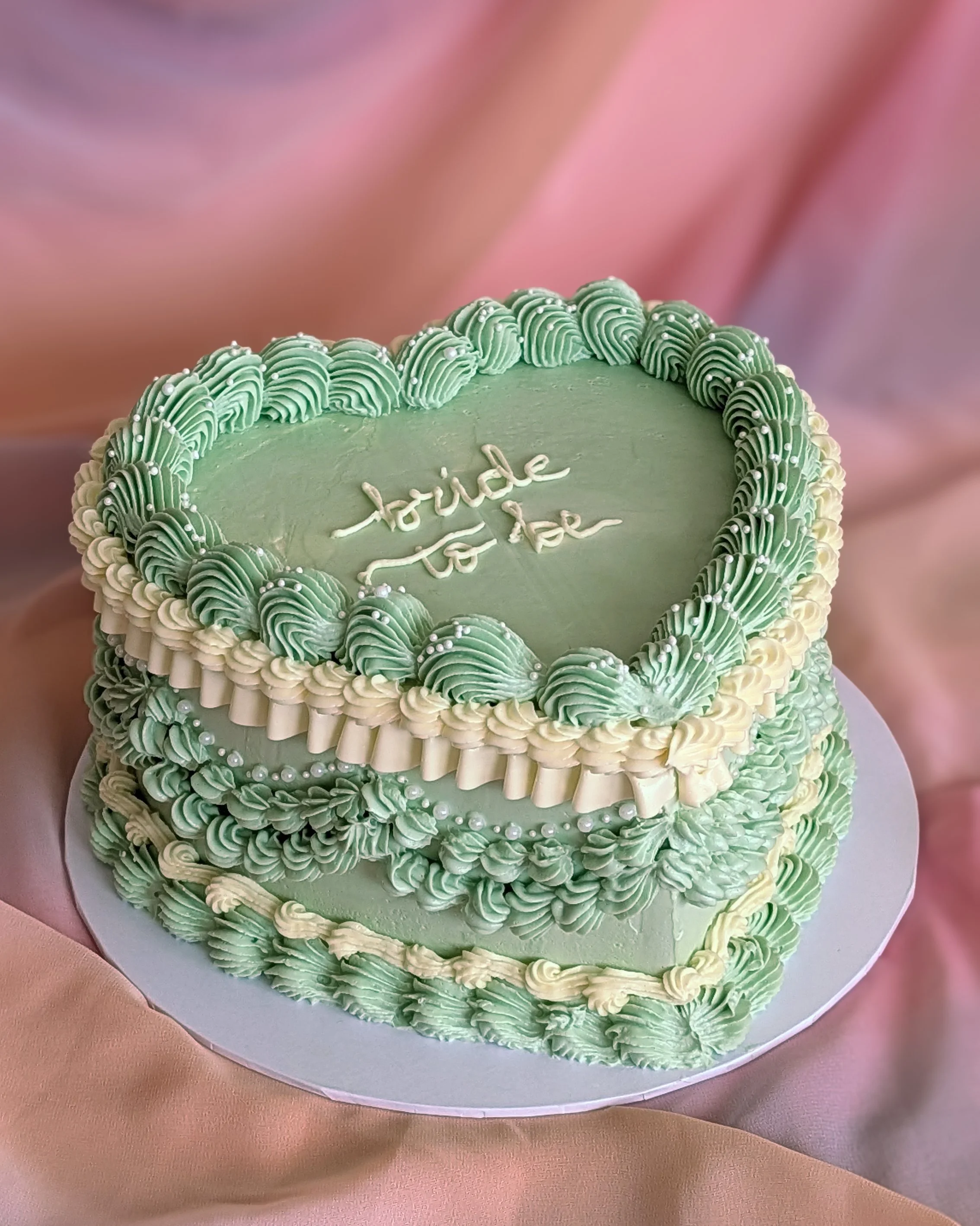 A round cake decorated with light green and white piping, and small silver edible beads. The cake has a message in white icing that says 'bride to be' and is placed on a white cake board on a pink and beige fabric background.