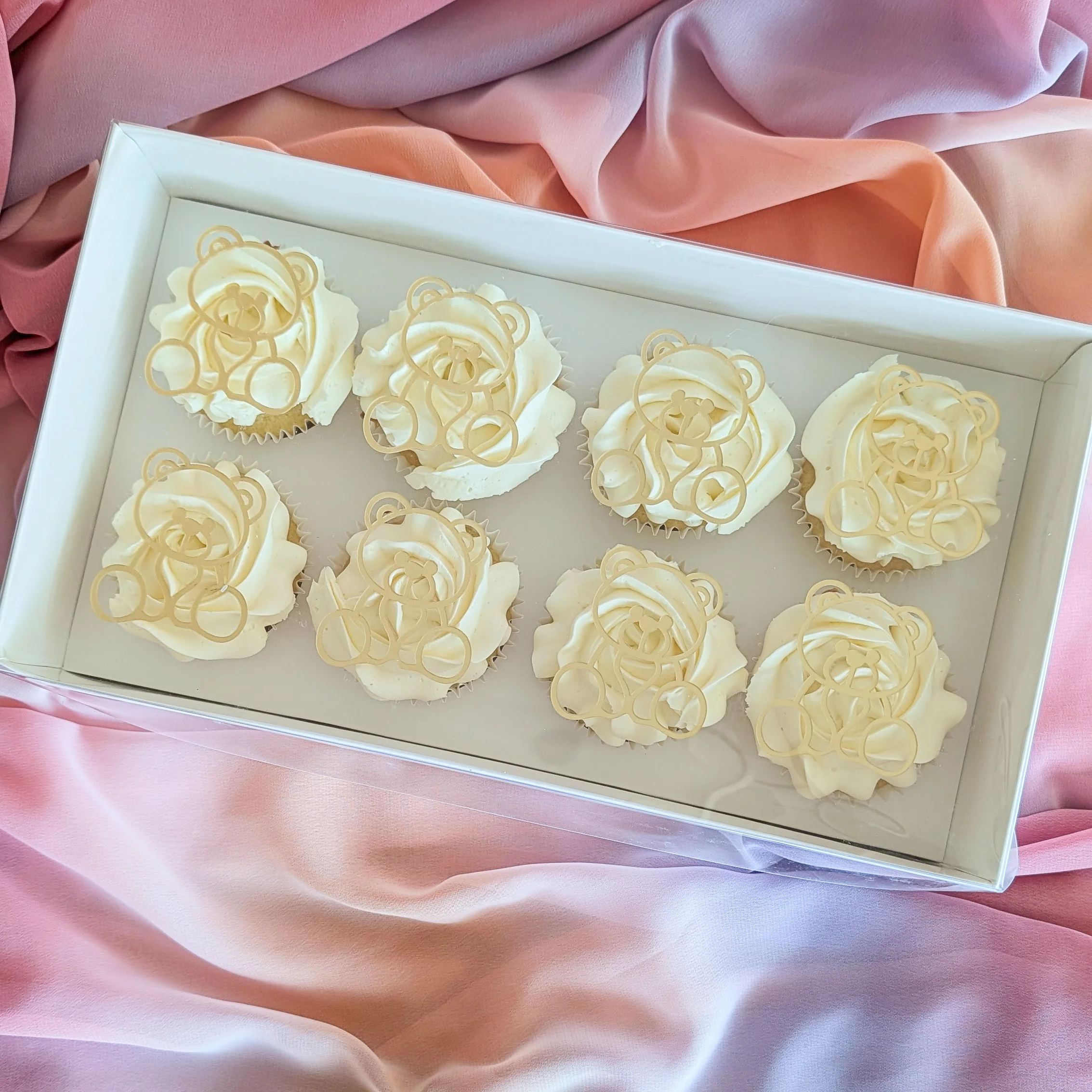 Box of cupcakes topped with white frosting and teddy bear-shaped white chocolate decorations, placed on pink and peach colored fabric.