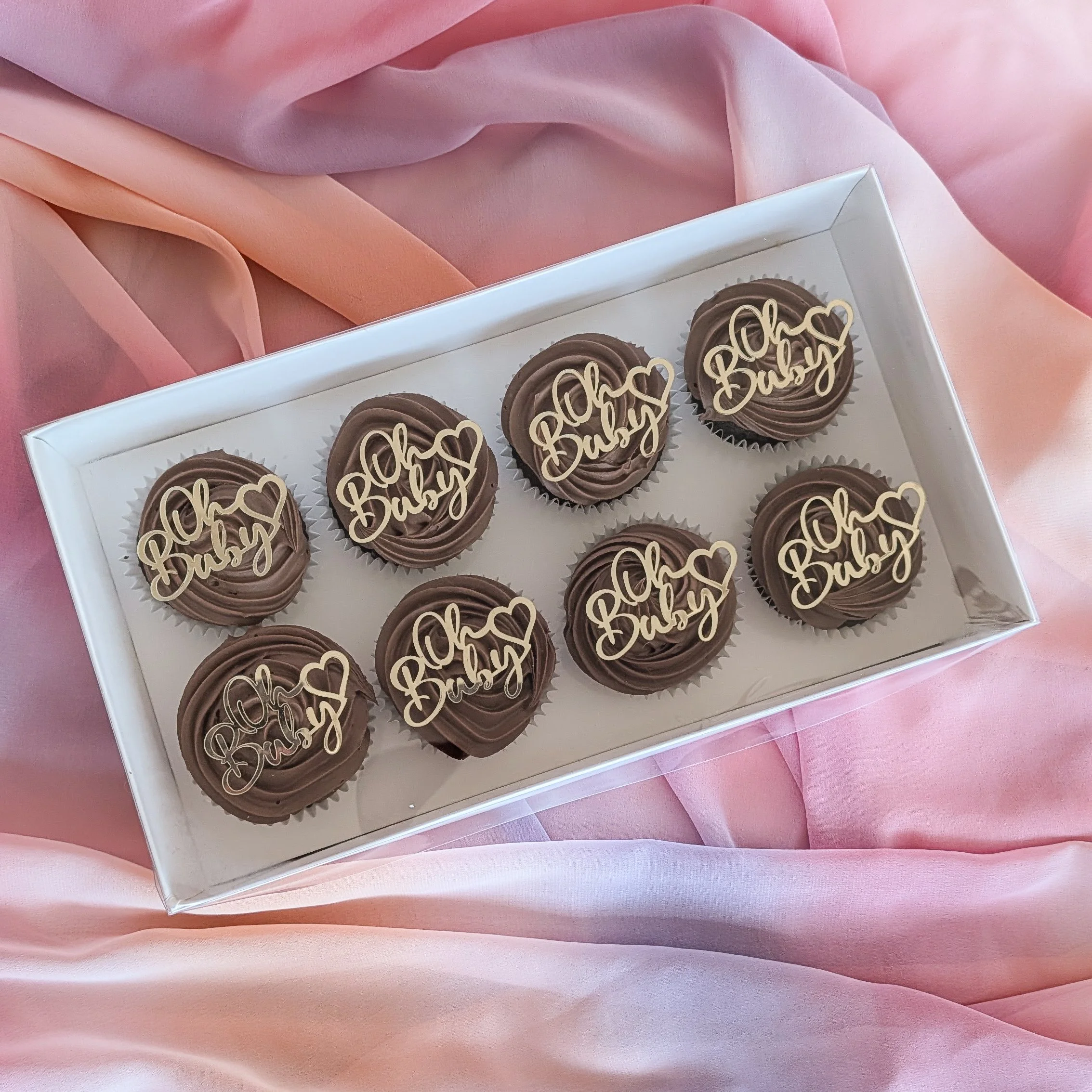 A white box contains several chocolate cupcakes decorated with chocolate swirls and edible gold-colored words that spell 'Baby' with hearts on top, placed on pink satin fabric.
