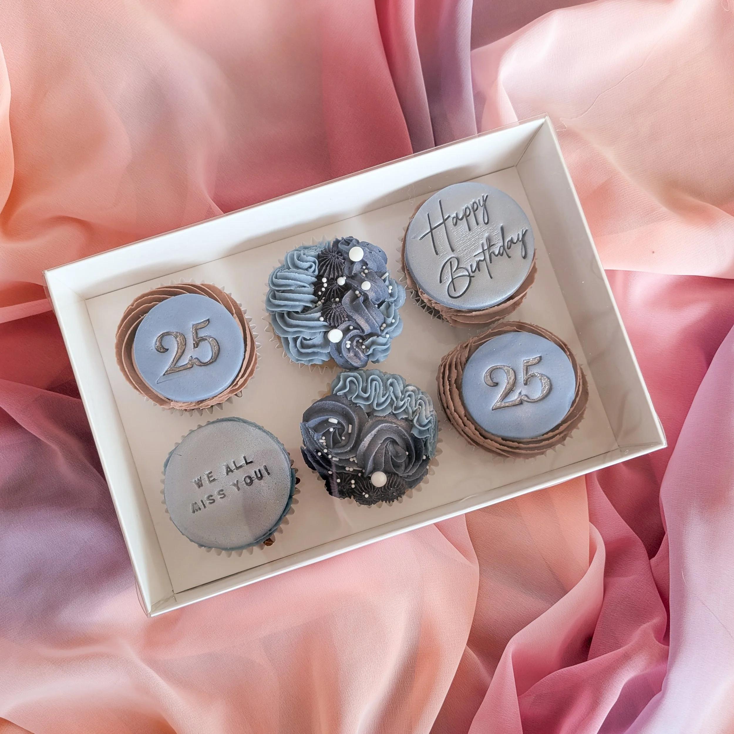 Box of decorated cupcakes with birthday messages, including one with a gray top and black writing that says 'Happy Birthday', set against pink fabric background.
