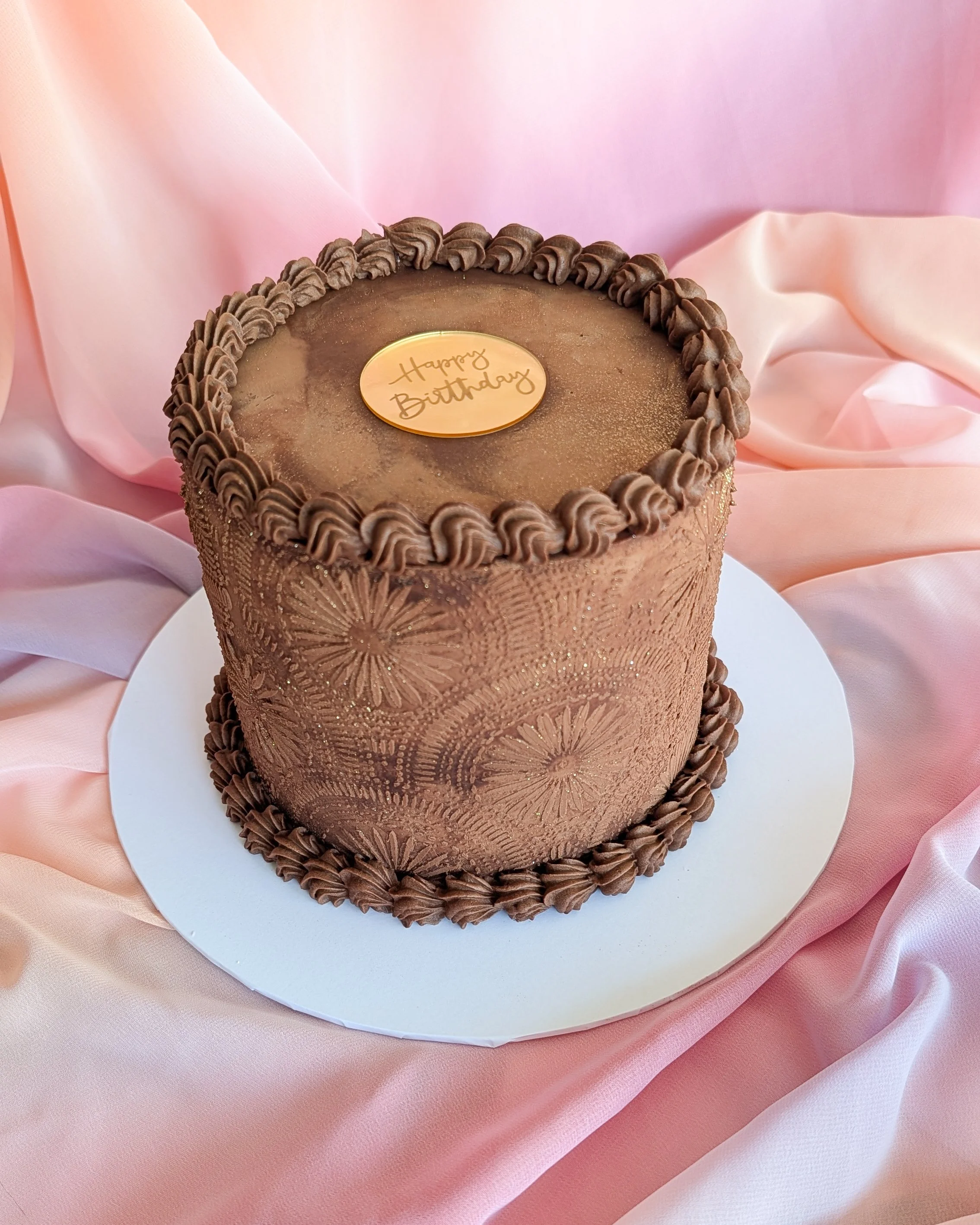 A round chocolate birthday cake with a gold "Happy Birthday" plaque on top, decorated with piped chocolate frosting around the edges, set on a white cake board on pink satin fabric.