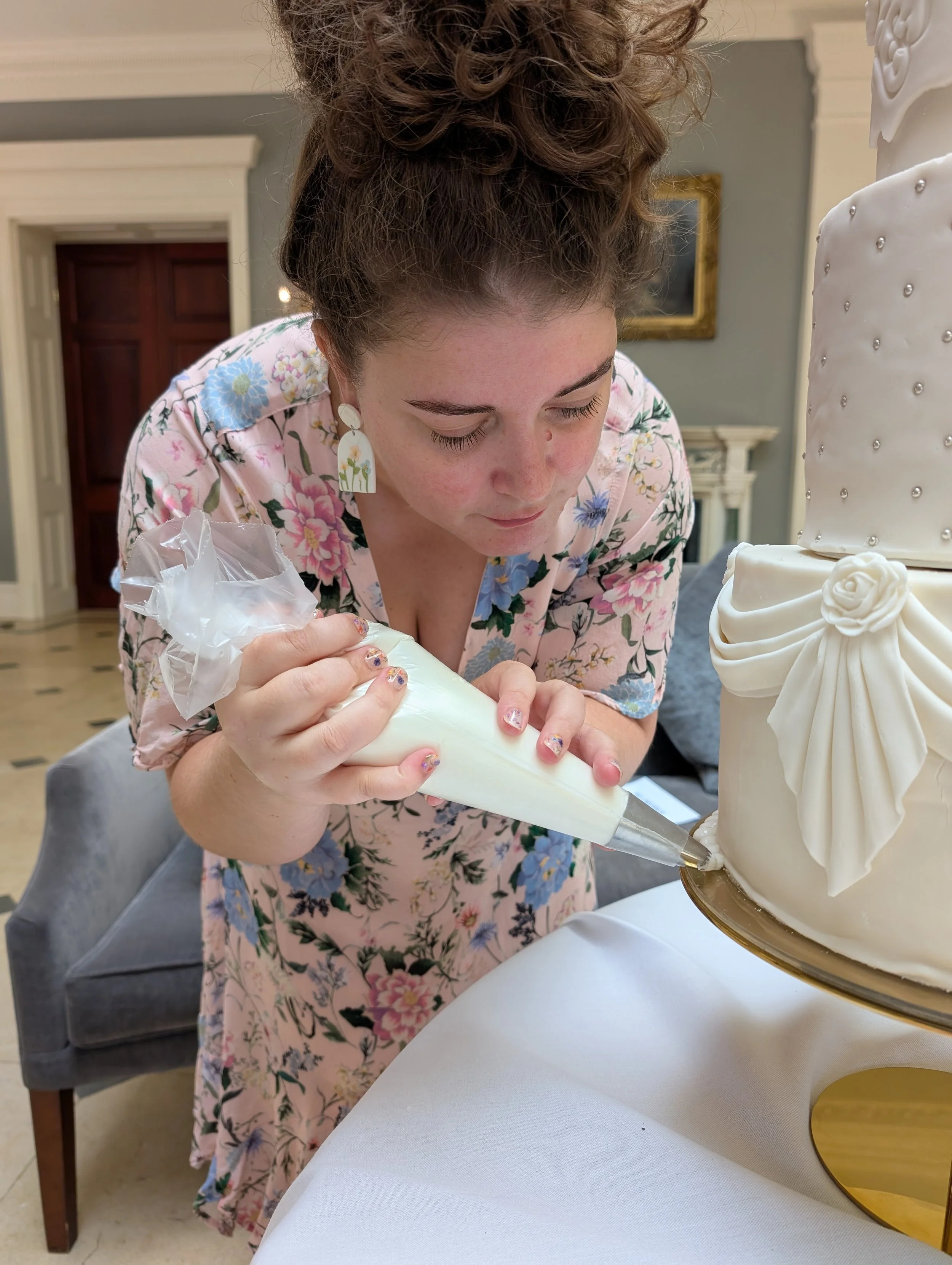 A woman decorating a white, multi-tiered cake with floral decoration and metallic embellishments, using a piping bag, in a decorated room.