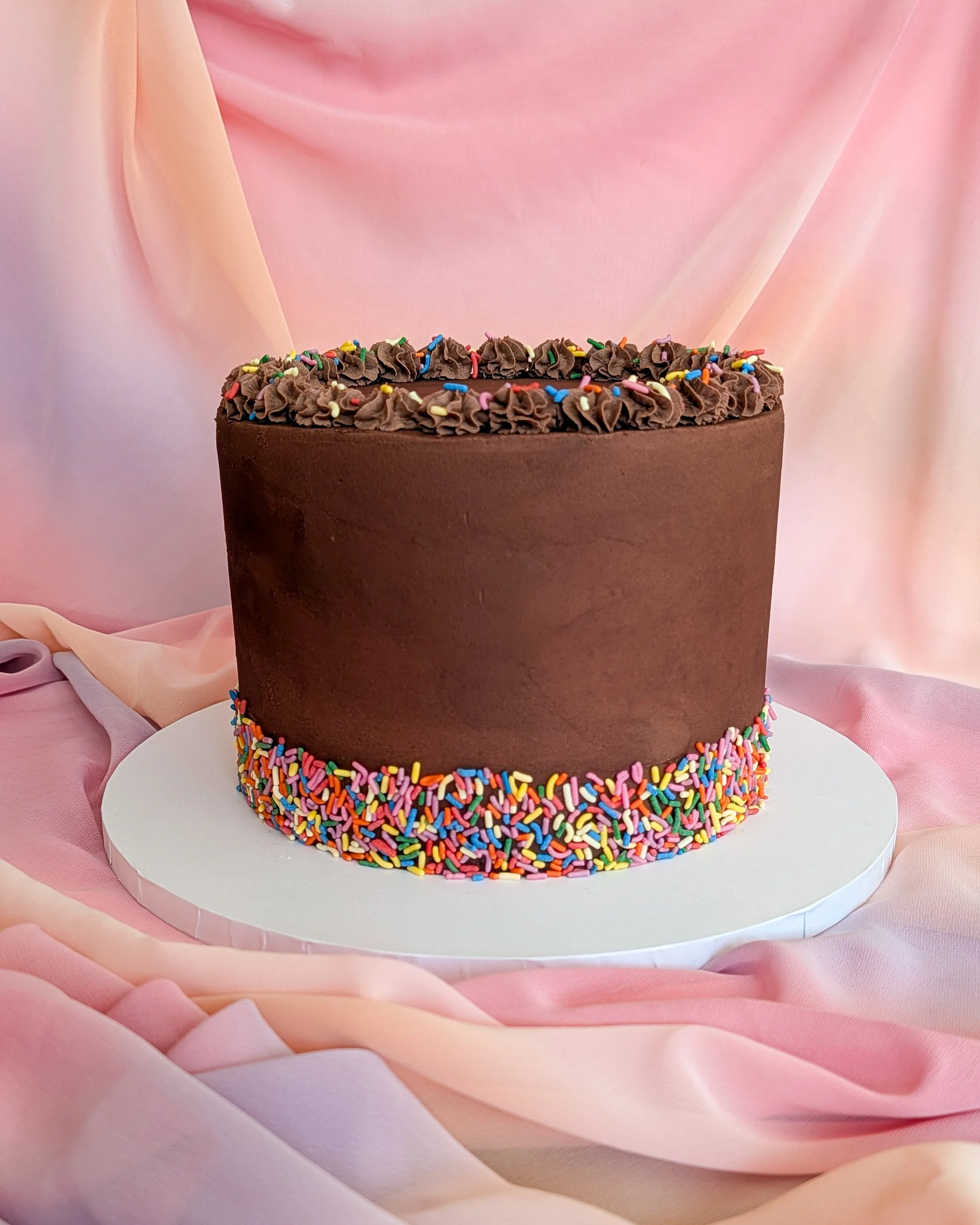 Chocolate cake with colorful sprinkles on top and around the base, set against pink satin fabric.