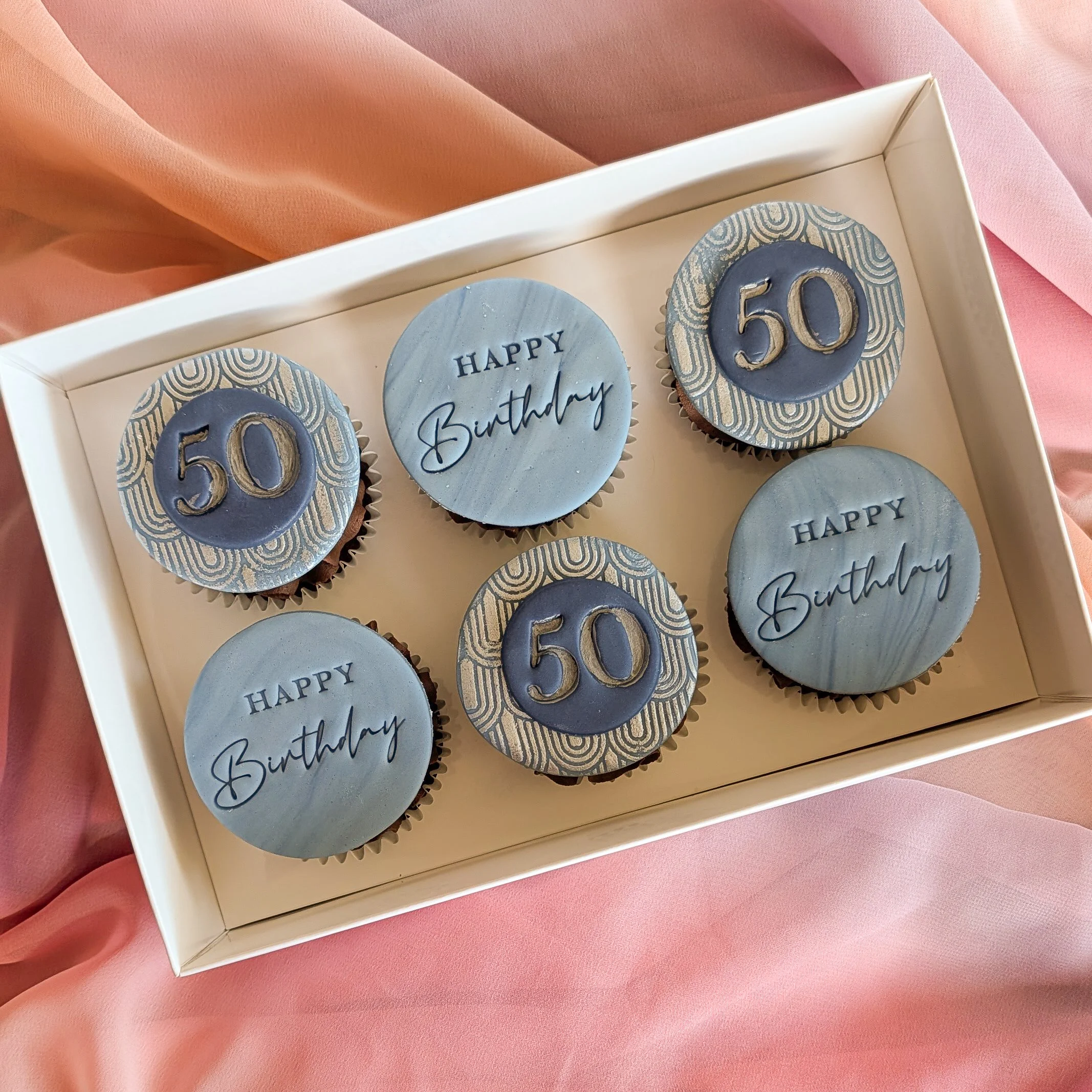 Celebratory cupcakes with blue and gold decorations, with plaques saying "Happy Birthday" and "50", in a white box on a pink satin background.