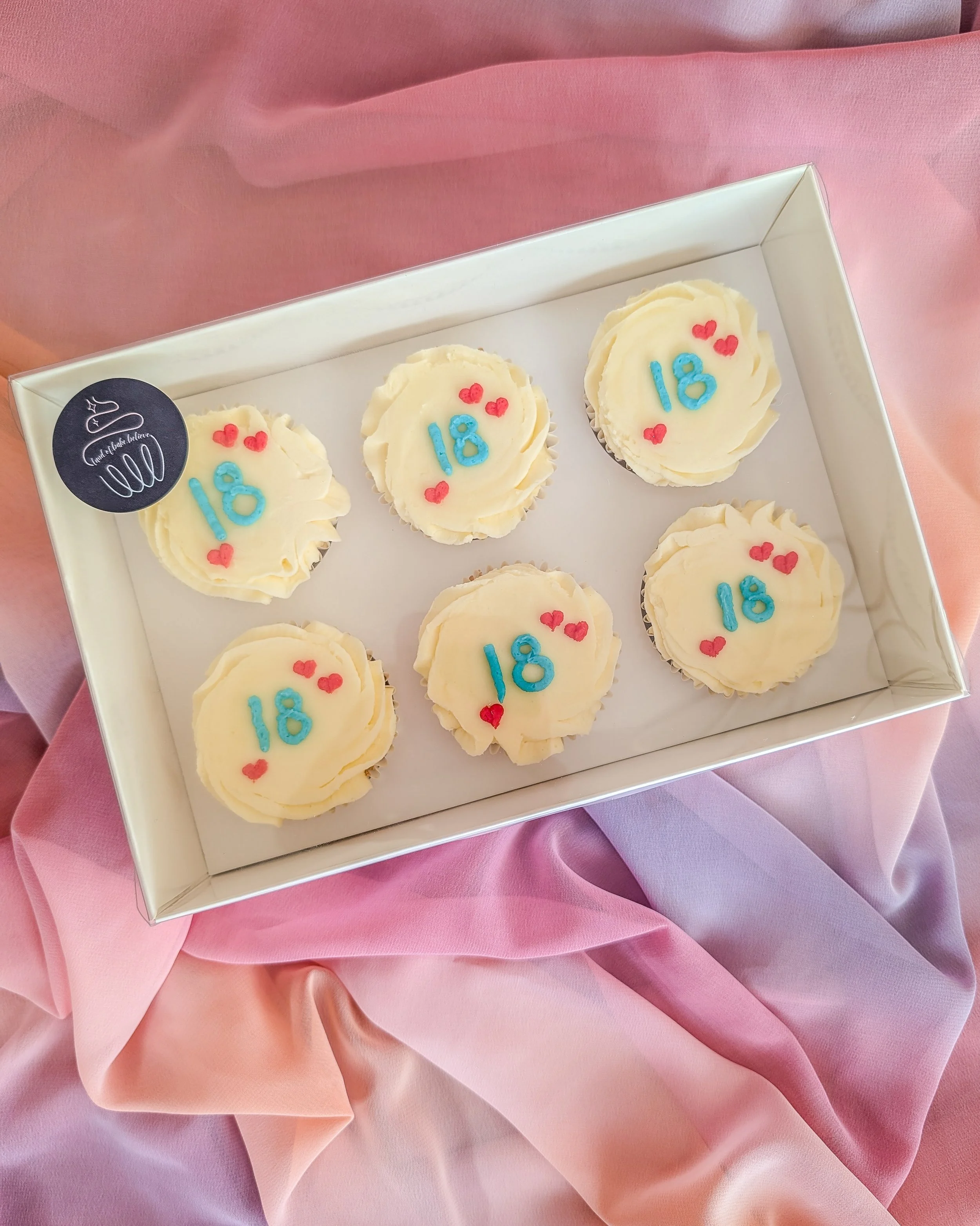Box of six cupcakes decorated with white frosting, each with a blue '18' and small red hearts, placed on pink fabric.