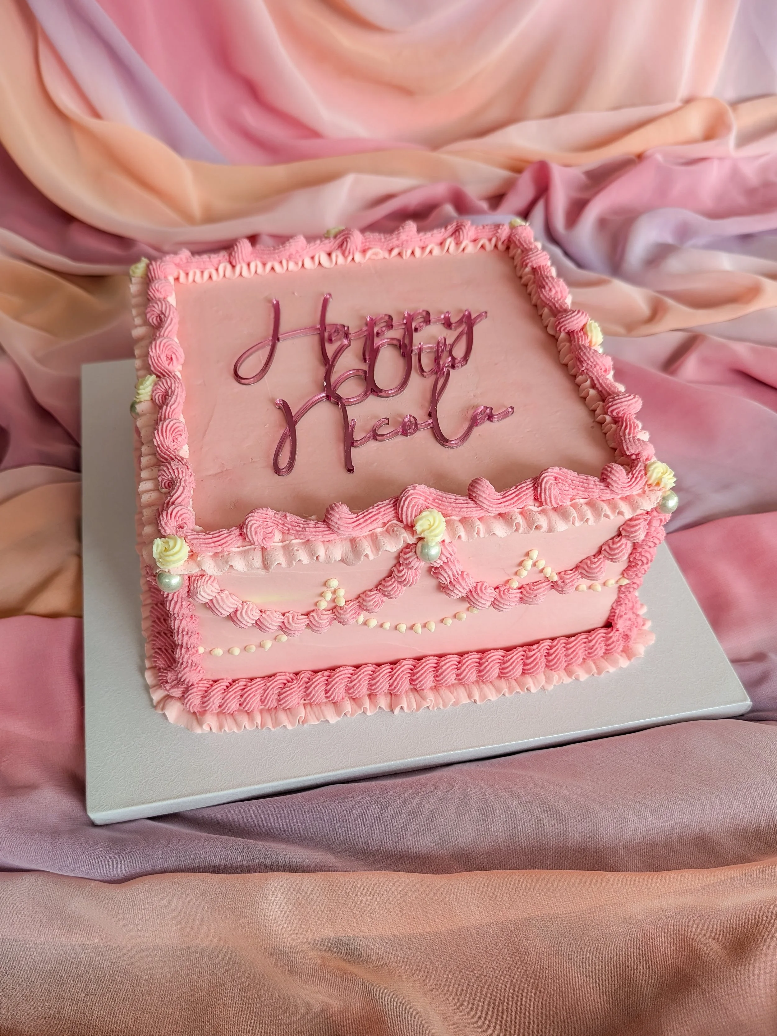 A pink birthday cake with pink icing and decorative border on white cake board, with the words 'happy birthday' written in pink cursive icing, placed on pink satin fabric.