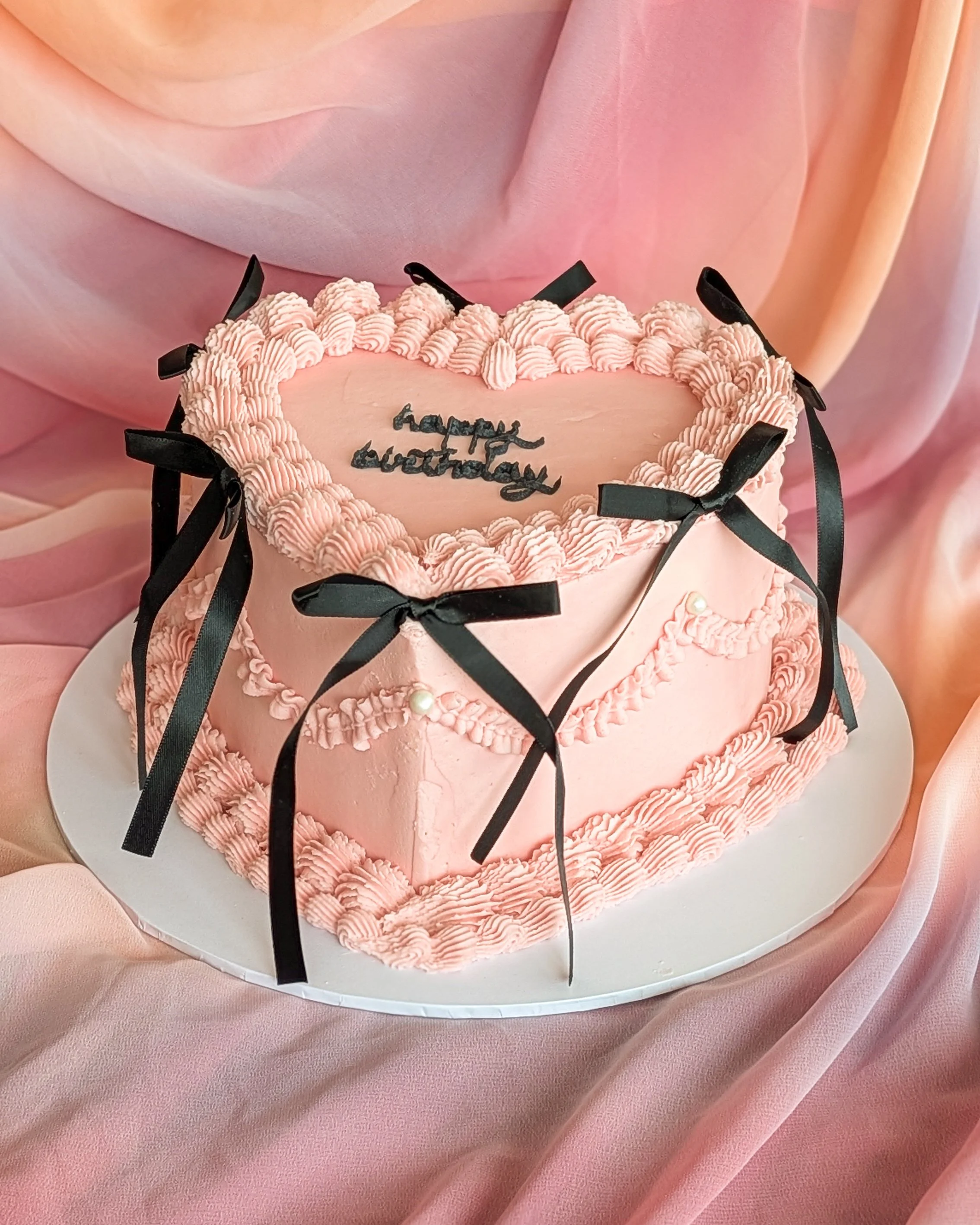 Pink heart-shaped birthday cake with ruffled pink icing, black ribbon accents, a single pearl decoration, and written message 'happy birthday' in black icing on top, with a pink fabric background.