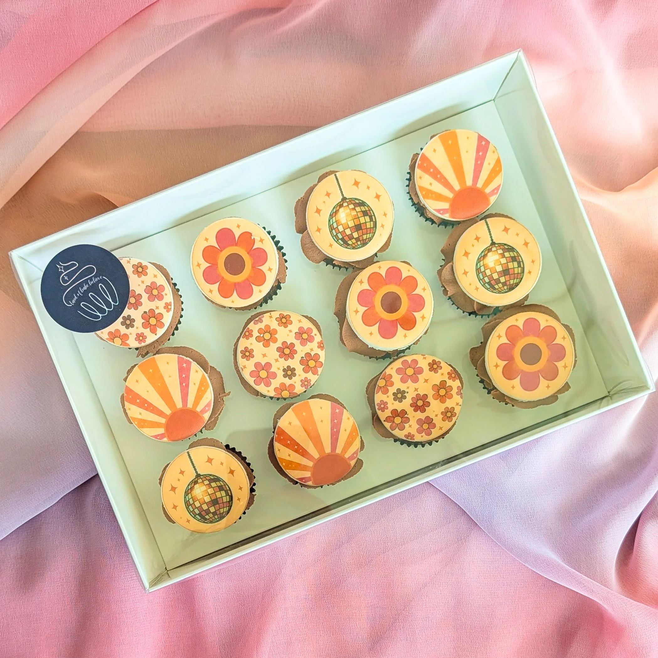 A box of cupcakes with colorful disco-themed decorations on top, placed on a pink and peach-colored fabric background.