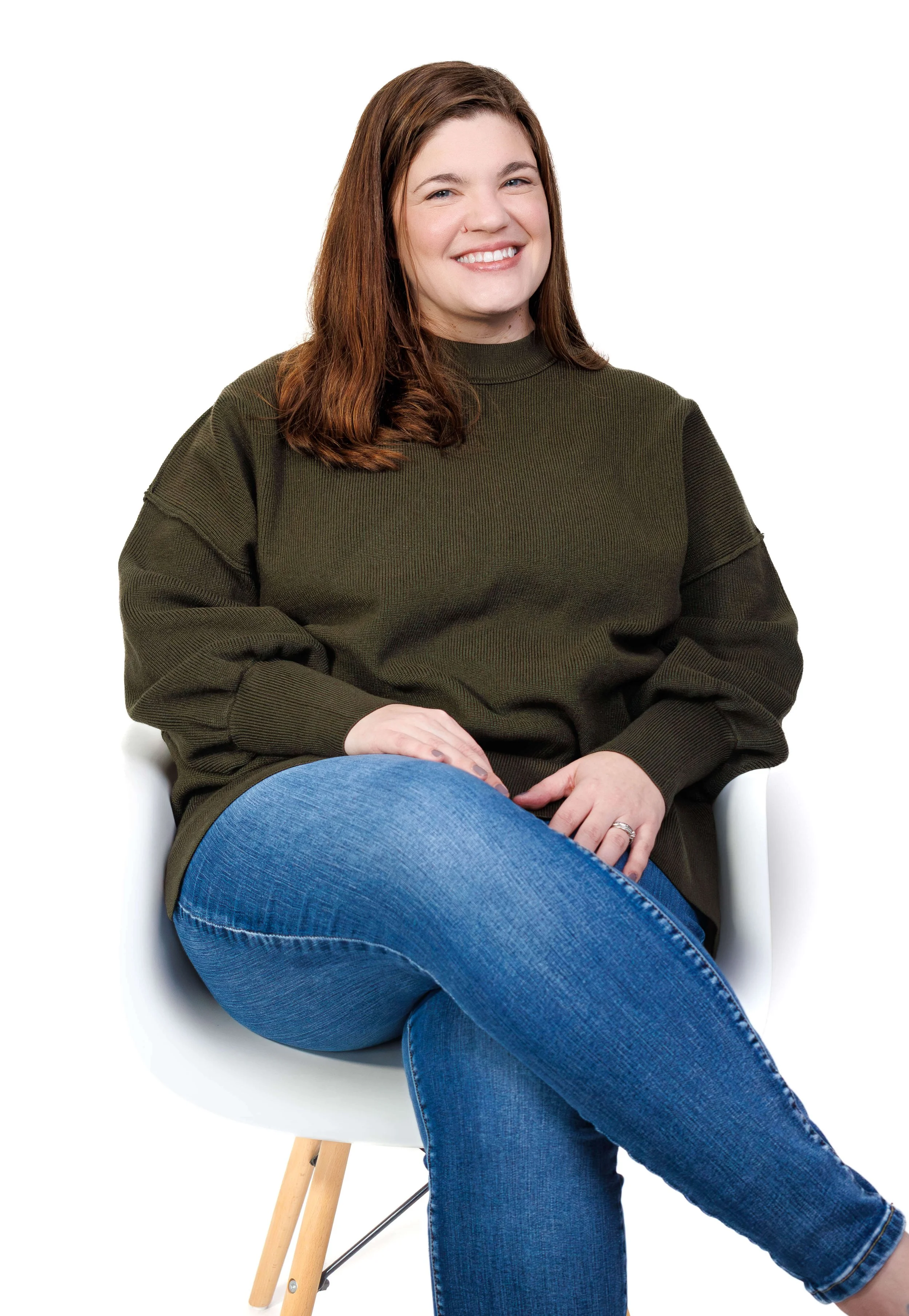 Sarah sits on a white chair in front of a white background. She is wearing a green sweater and jeans.