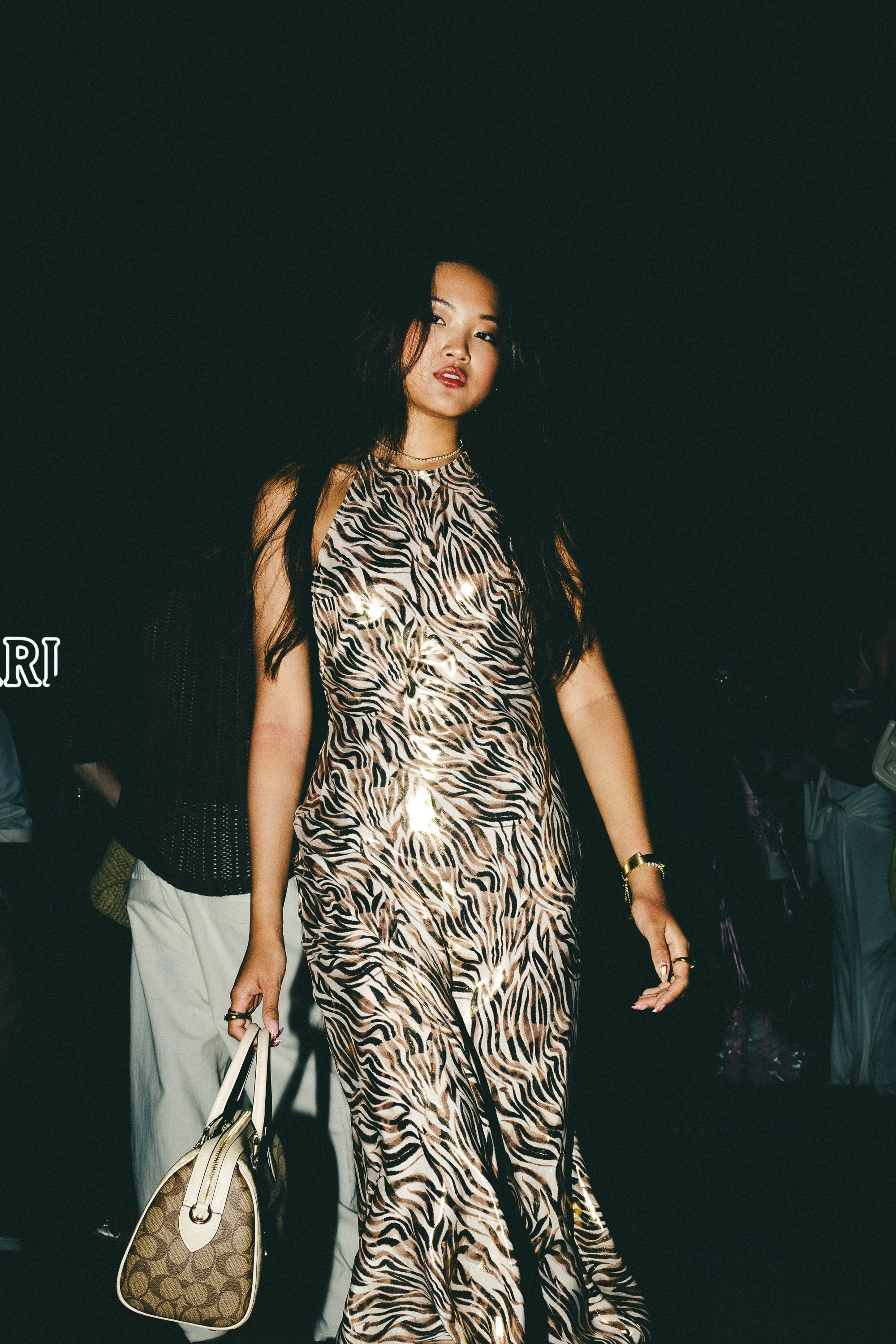 A woman wearing a sleeveless animal print dress with gold and black patterns, holding a designer handbag, walking in a dark setting with other people around.
