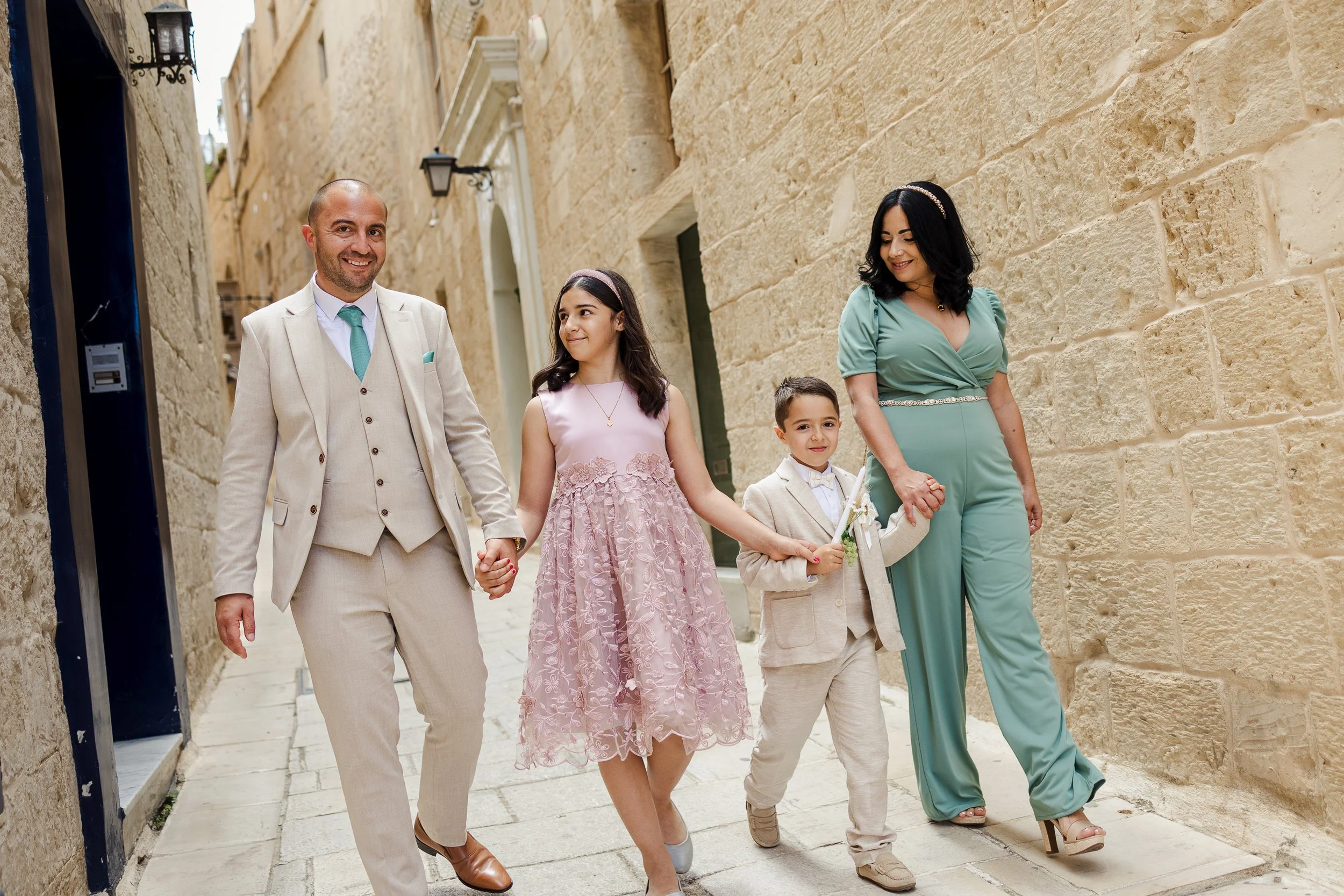 Family of five walking in a narrow street with stone walls, dressed in semi-formal attire, holding hands, smiling, during daytime.