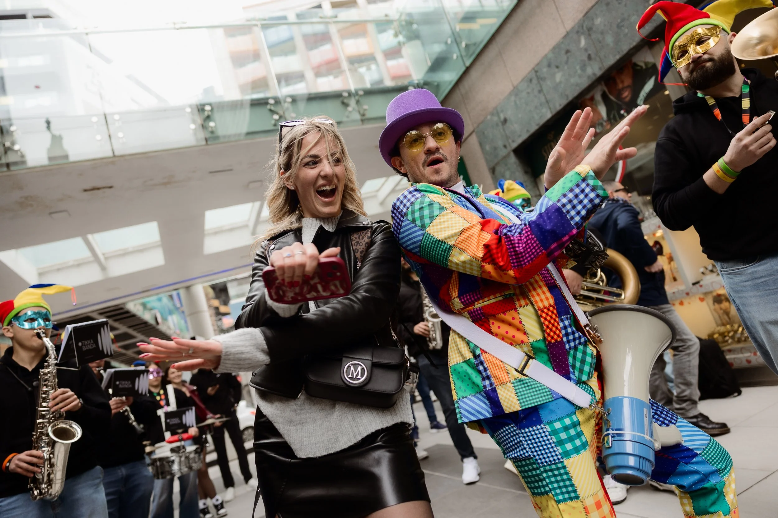 People in colorful costumes enjoying a parade. One person is dressed in a patchwork suit with a musical instrument, and others are wearing masks and accessories.